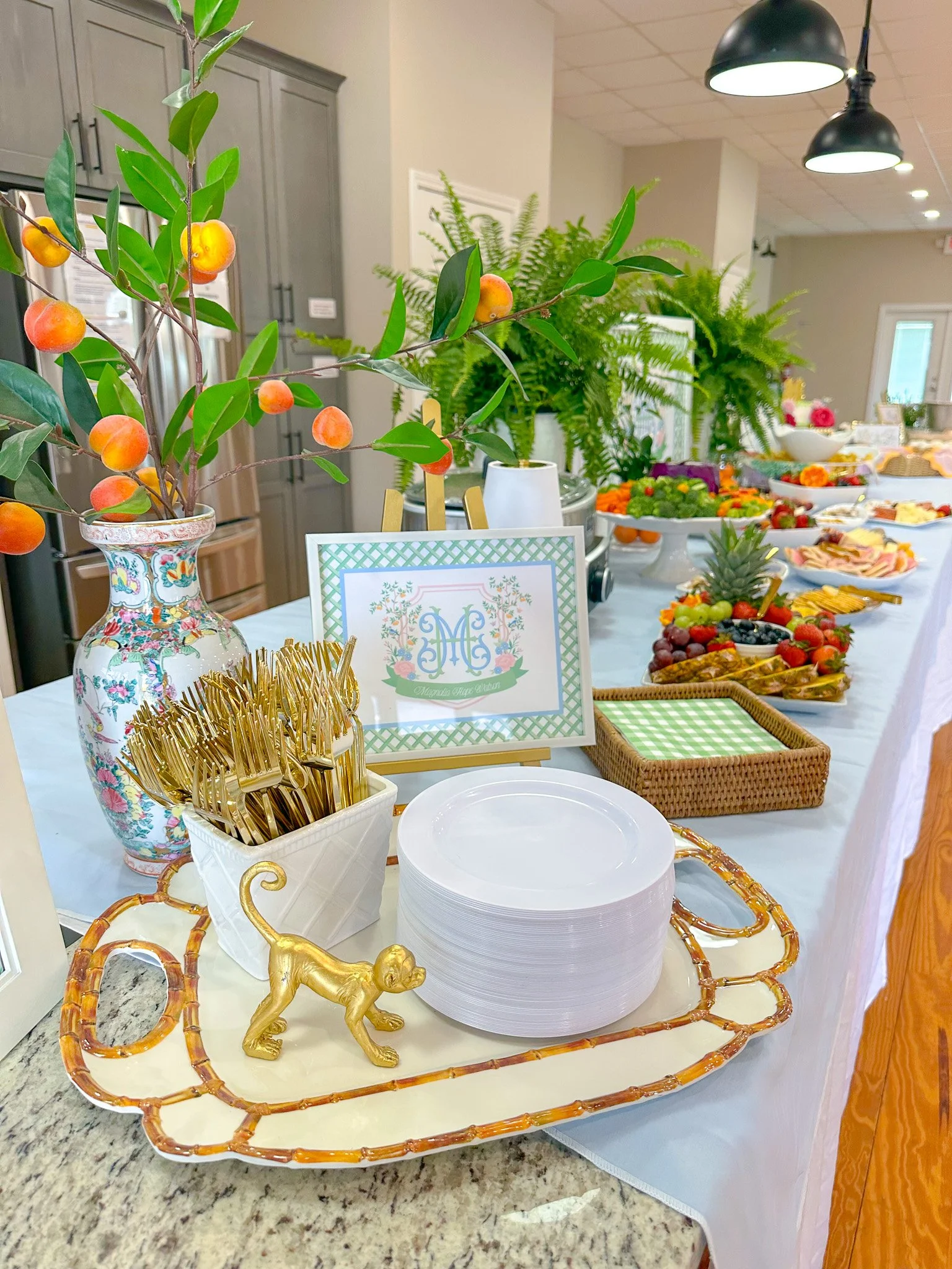 A decorated table with a branch of peach-colored fruit and green leaves in a floral vase, gold utensils in a white container, a stack of white plates, and a boxed gold monkey figurine. The background shows a buffet of various foods, greenery, and a s