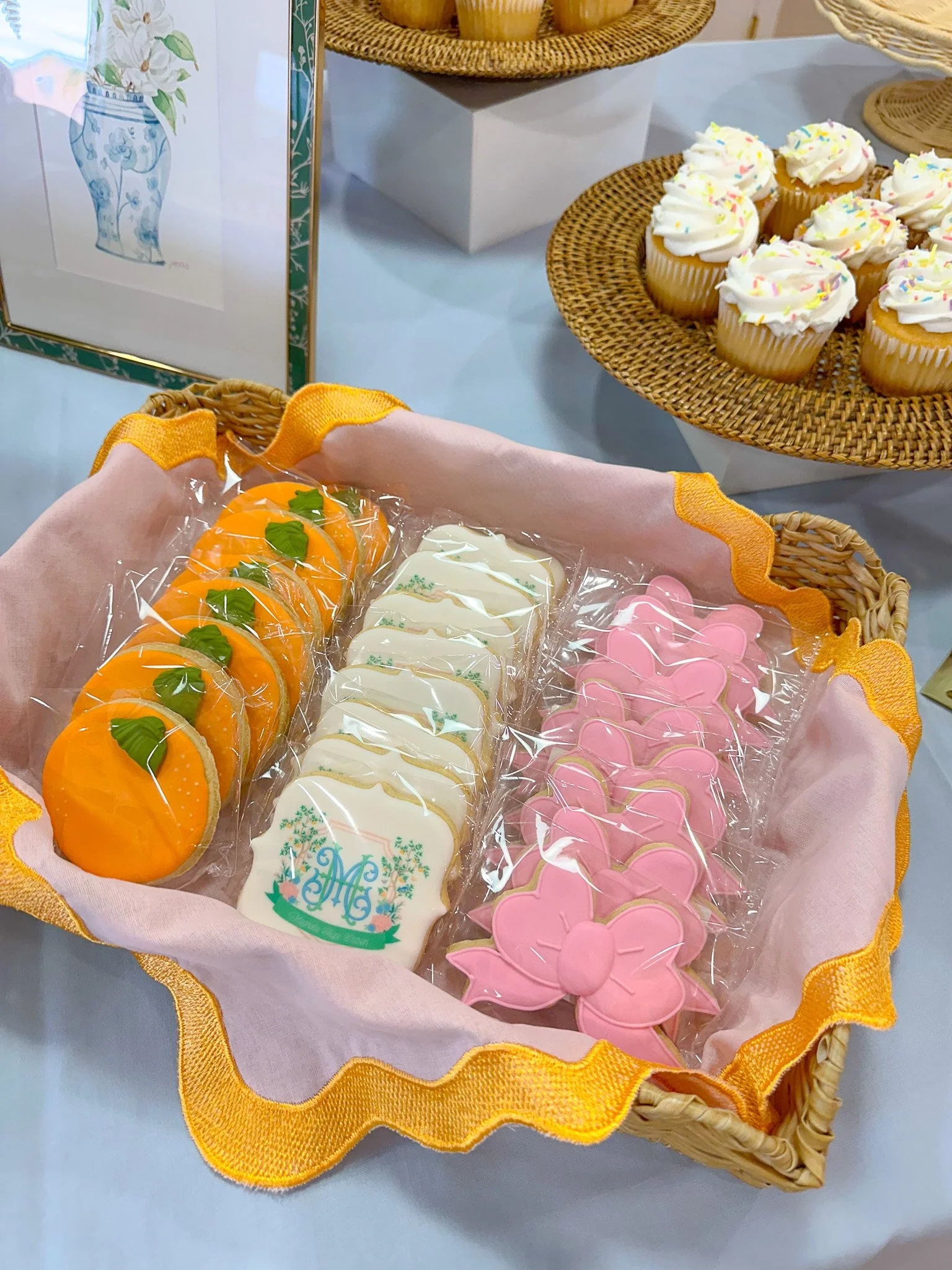 A basket filled with decorated cookies, including orange, white with green designs, and pink butterfly-shaped cookies, on a table with cupcakes decorated with frosting and sprinkles in the background.