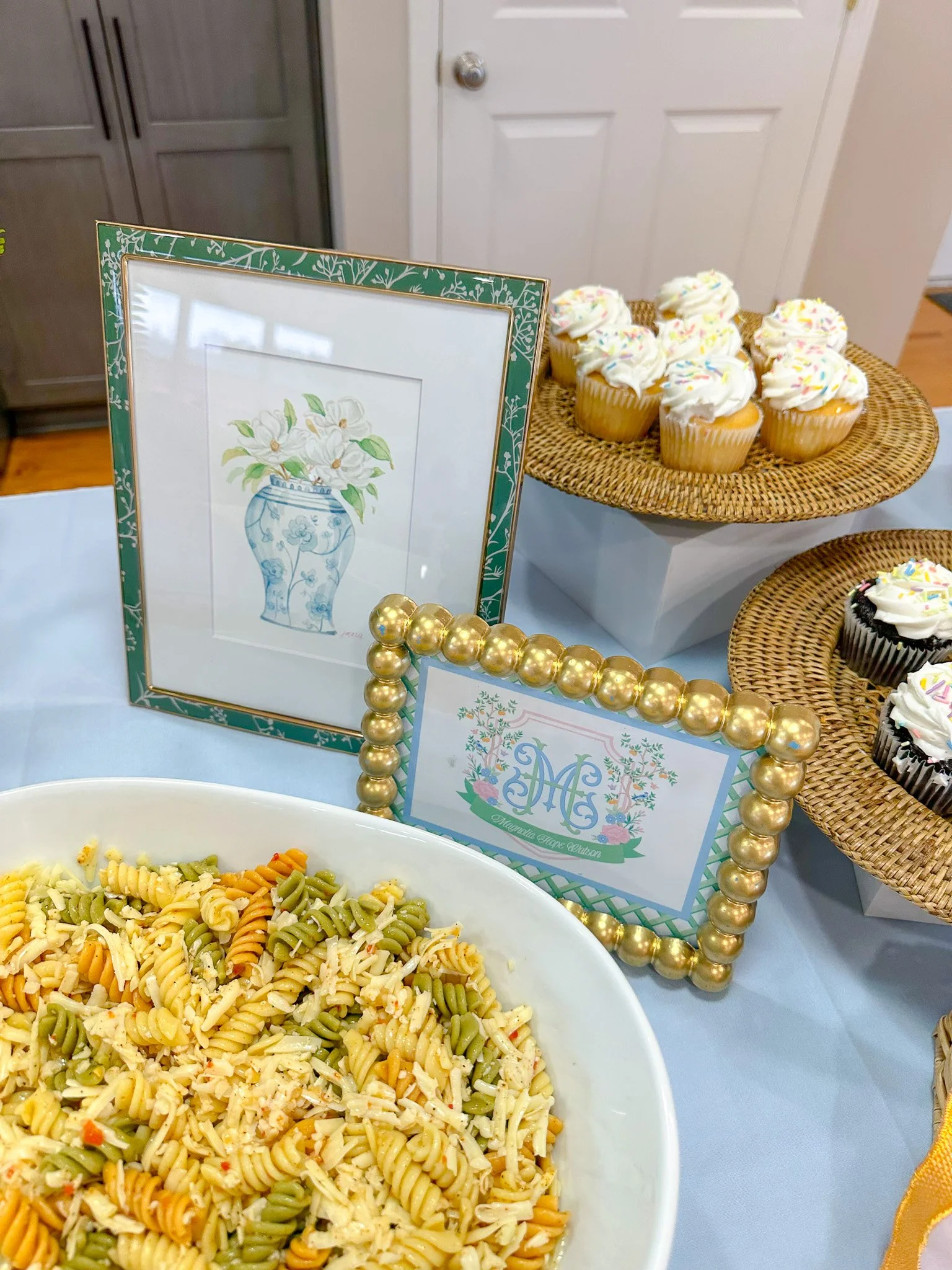 Dessert table with cupcakes, pasta salad, and decorative signs, including a framed artistic illustration of a vase with flowers.
