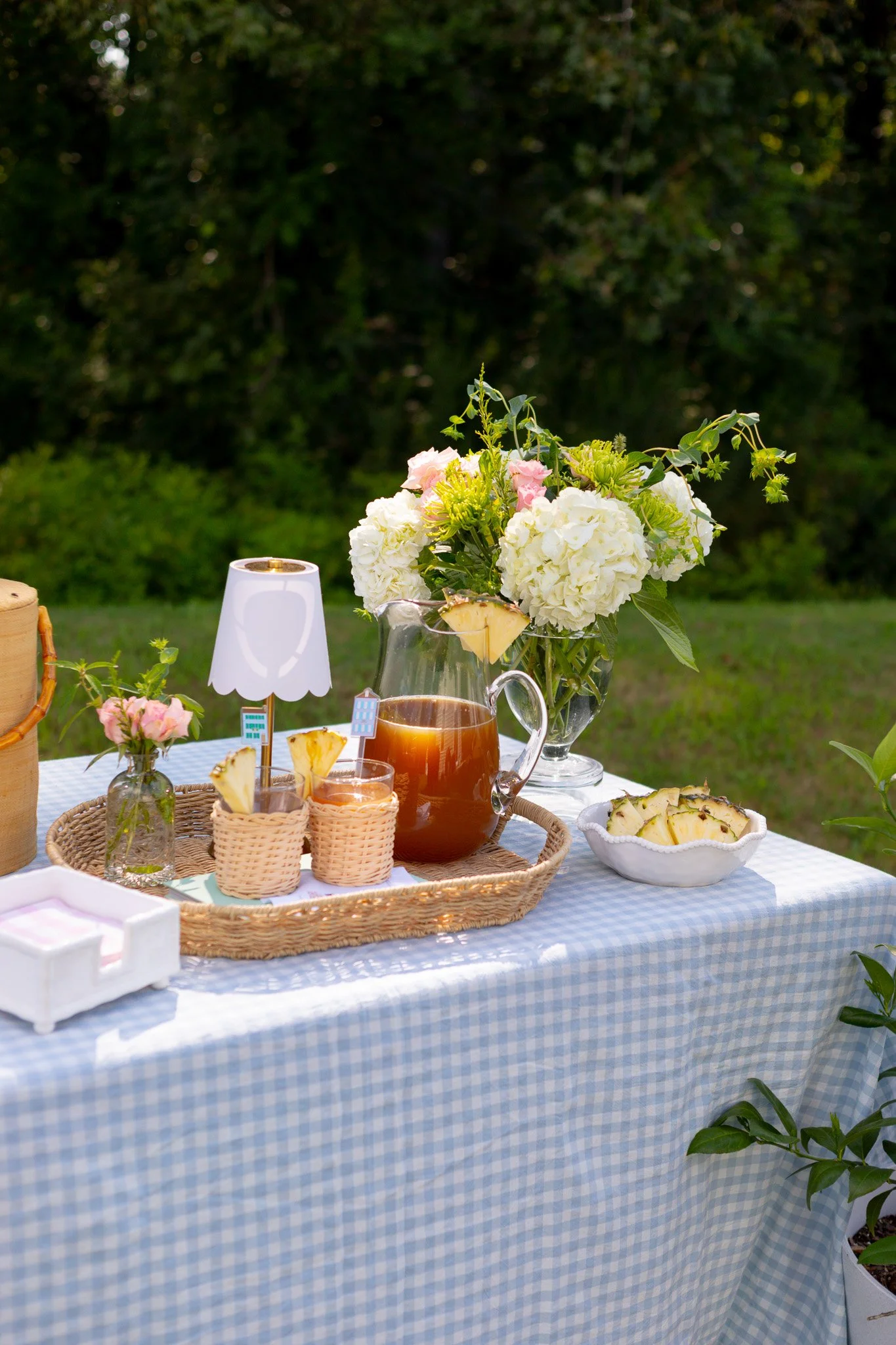 Outdoor table set for a meal with a blue checkered tablecloth, white flowers, pineapple slices, iced tea, and a small lamp, against a background of green trees.