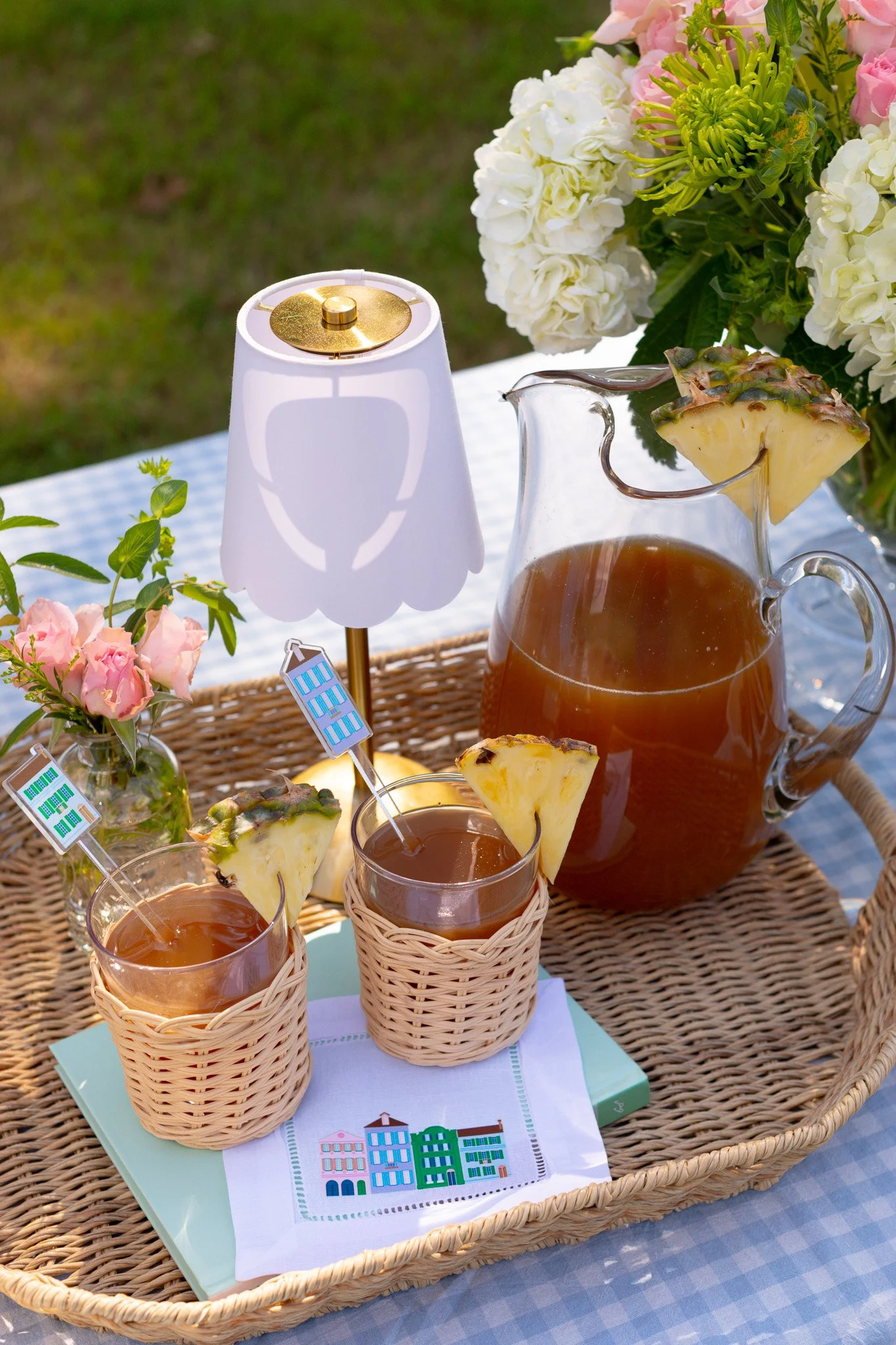 A wicker tray with two glasses of iced tea garnished with pineapple slices and small flags, a pitcher of iced tea with pineapple, a small white lamp, and pink and white flowers on a table with a checkered tablecloth outside.