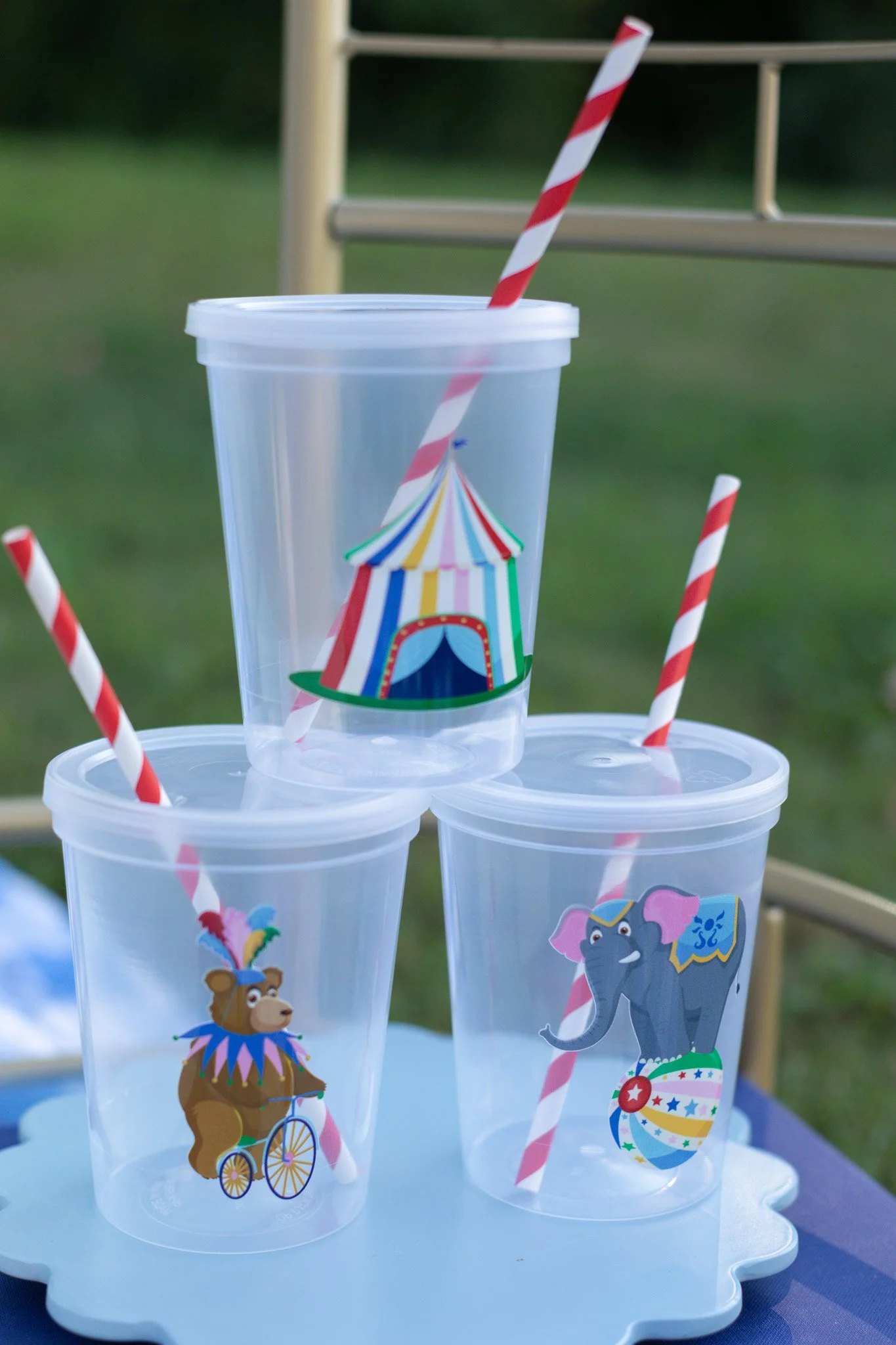 Three empty party plastic cups with colorful circus animal stickers and red and white striped straws on a light blue tray outdoors.