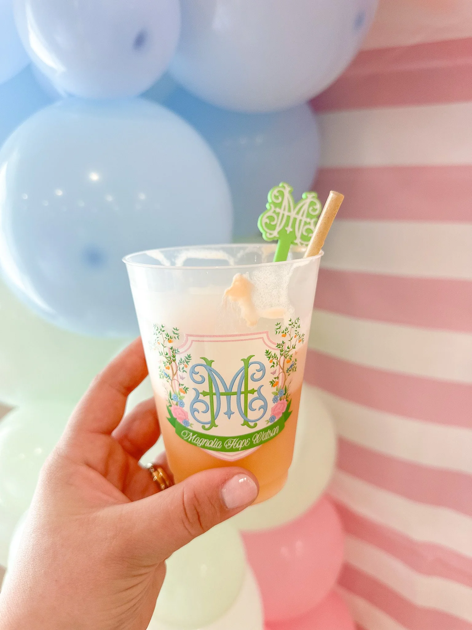 Close-up of a hand holding a clear plastic cup with a pink beverage, decorated with a monogram and floral design, in front of pastel-colored balloons and a pink and white striped background.