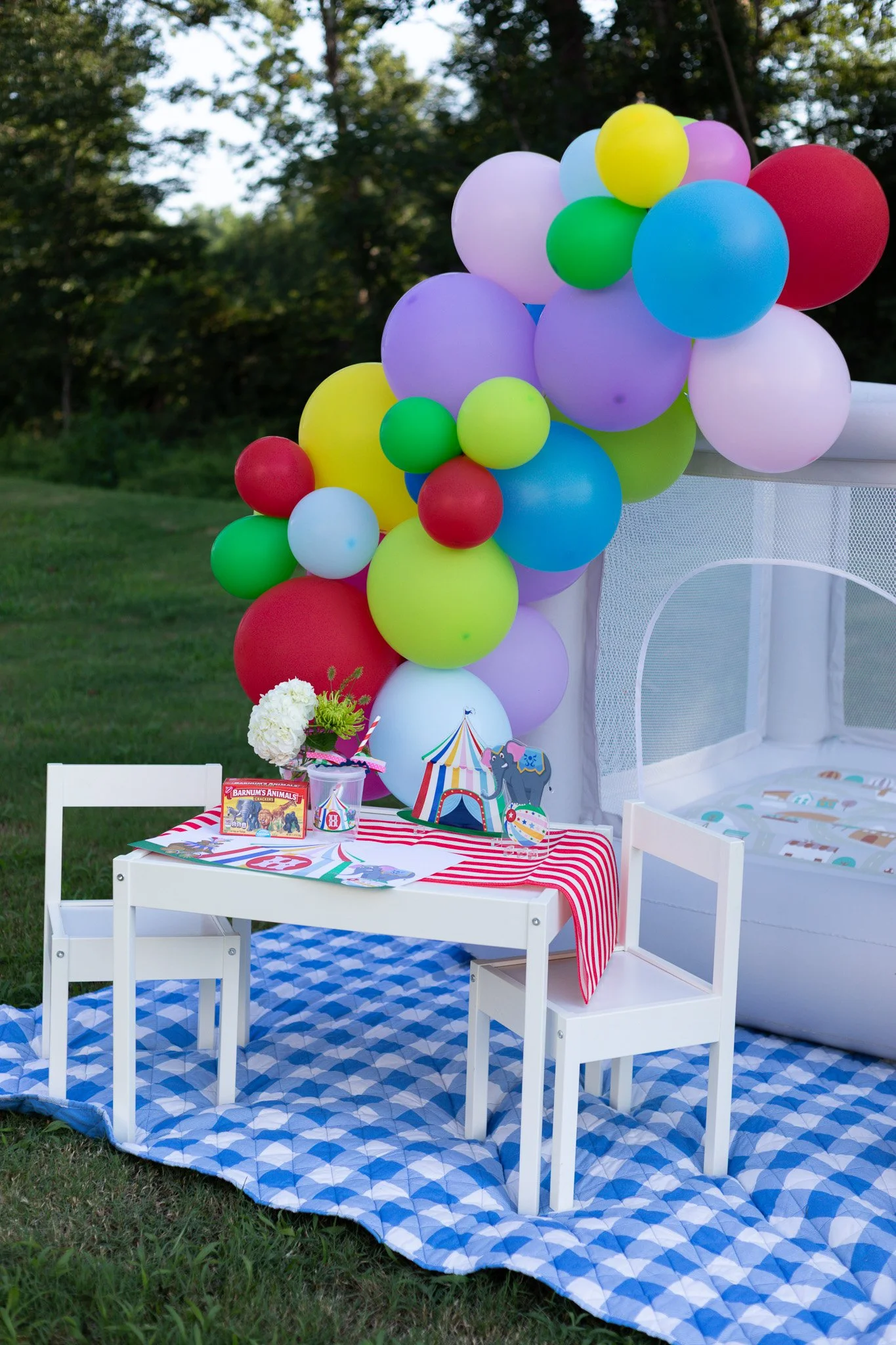 Colorful balloons tied to a small table with birthday decorations, set outdoors on a grassy area.