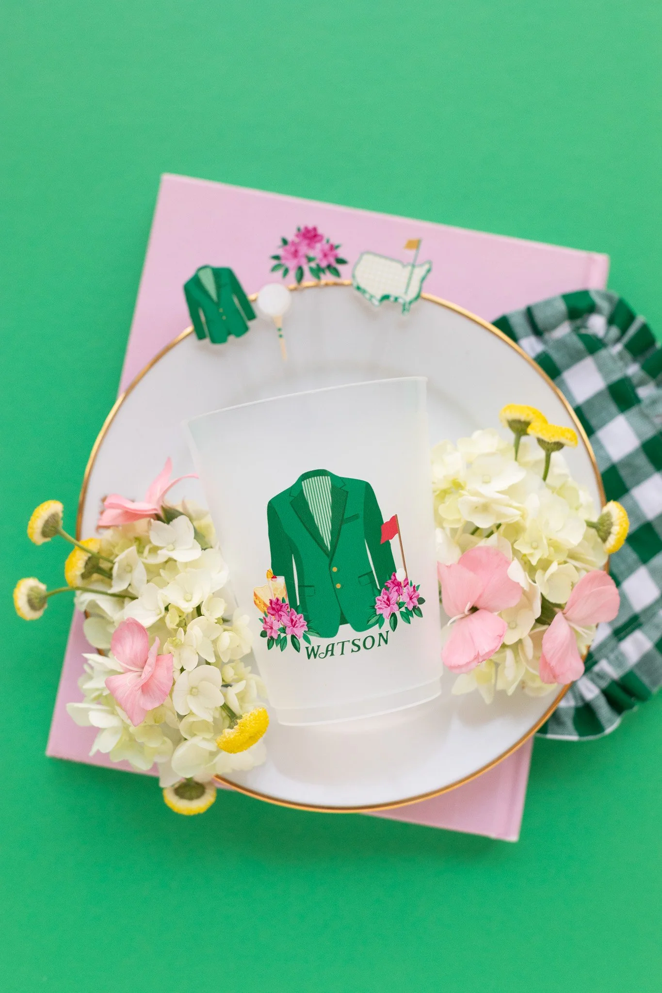 Decorative glass with a suit jacket design labeled 'WATSON,' surrounded by white and pink flowers on a white plate, on a pink and green background.
