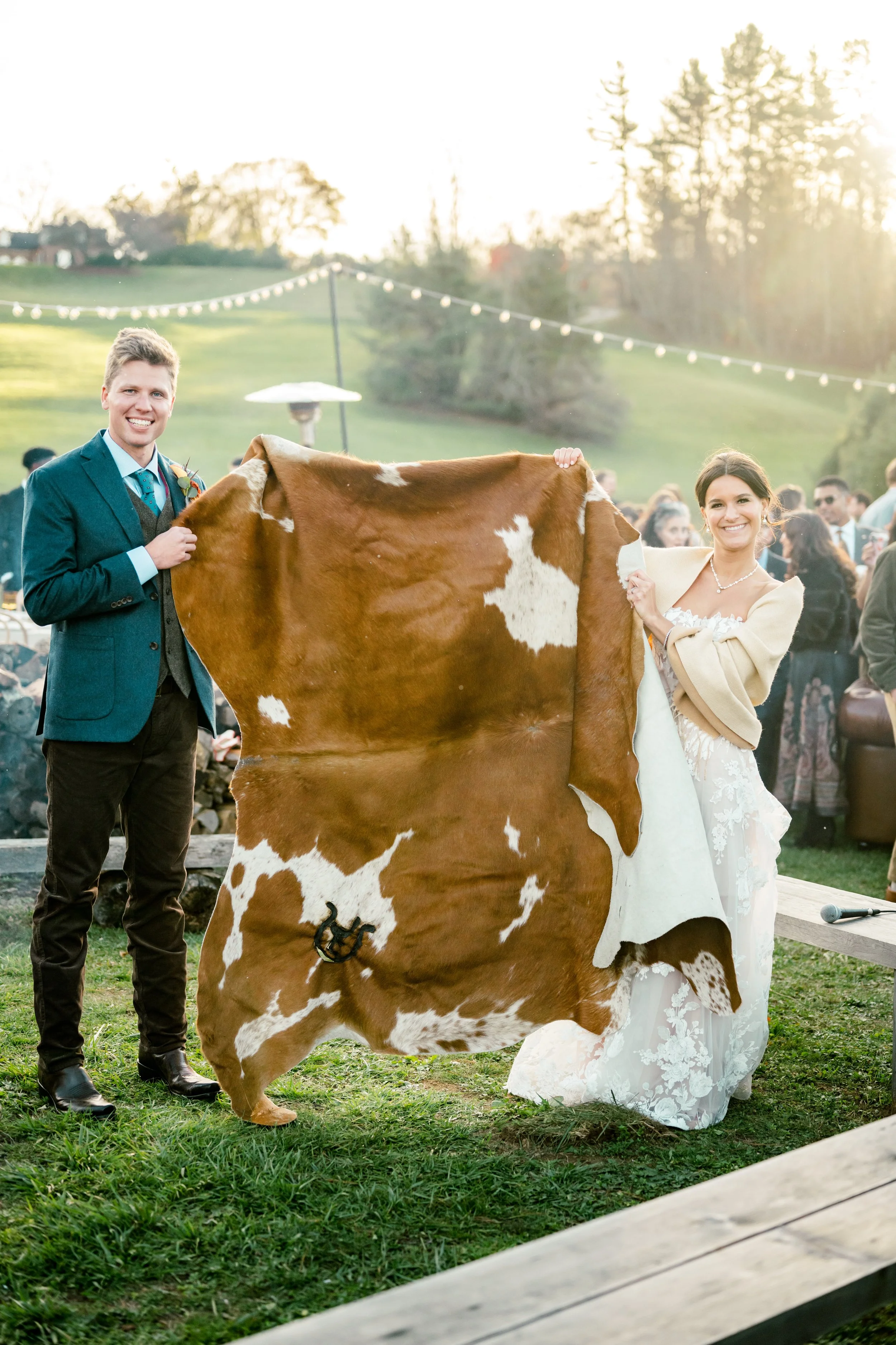 A bride and groom holding a cowhide rug at an outdoor wedding reception during sunset, with guests in the background.
