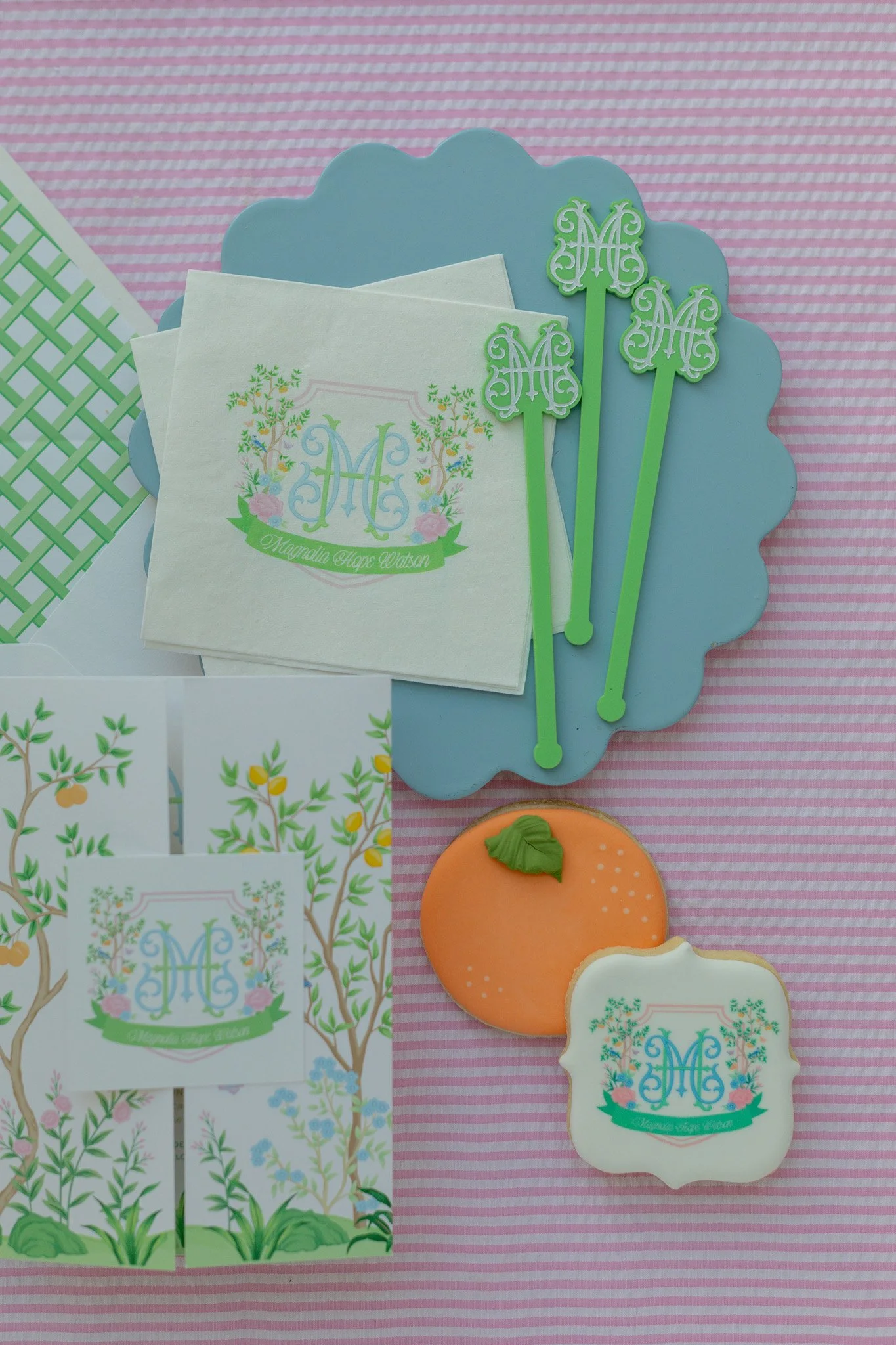 Party supplies with a garden theme, including monogrammed napkins, cake toppers, and cookies, with a pink and white striped background.