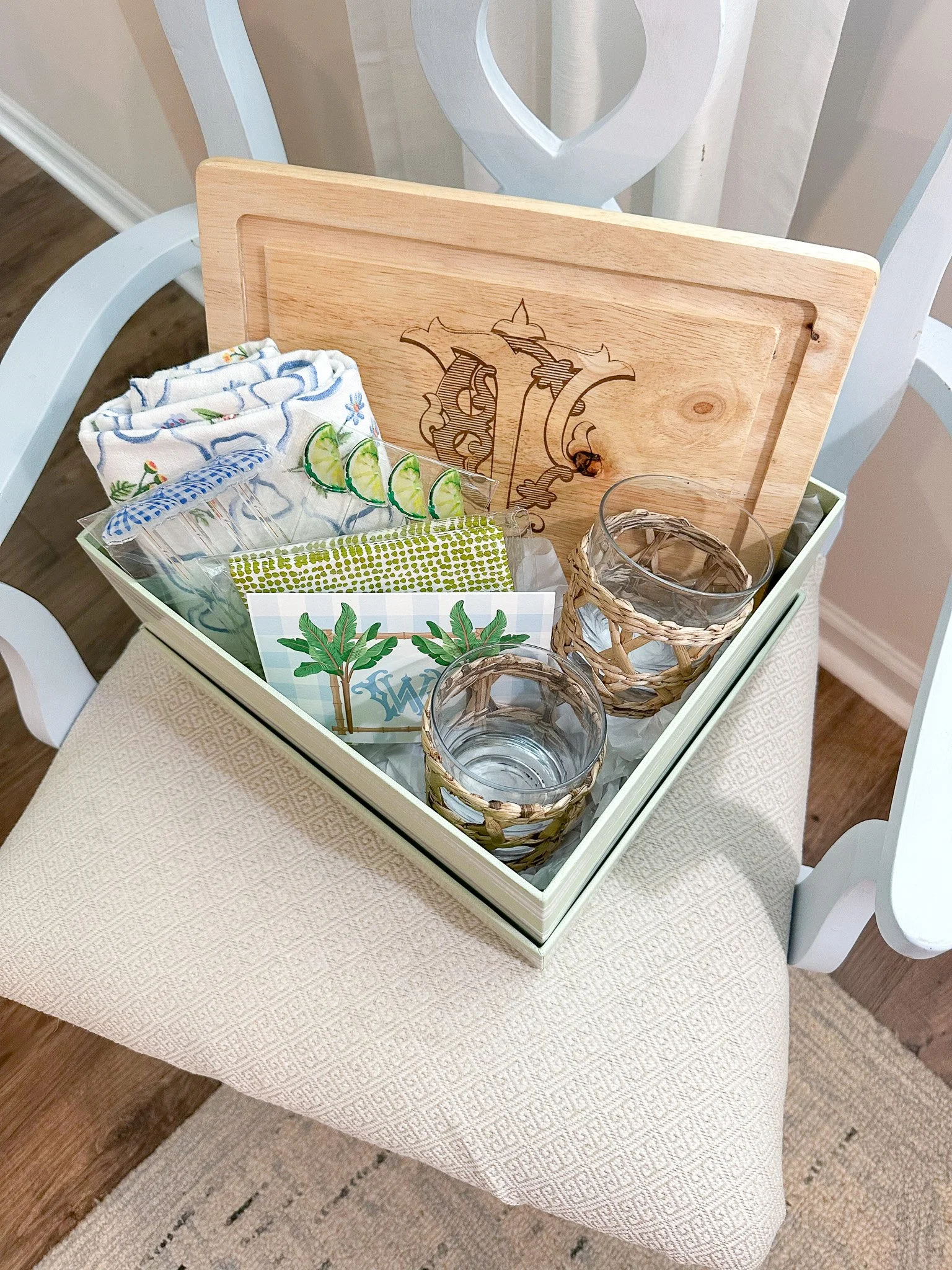 A gift box containing glasses with woven covers, lime slices, towels, napkins, and a wooden board inside a white chair.