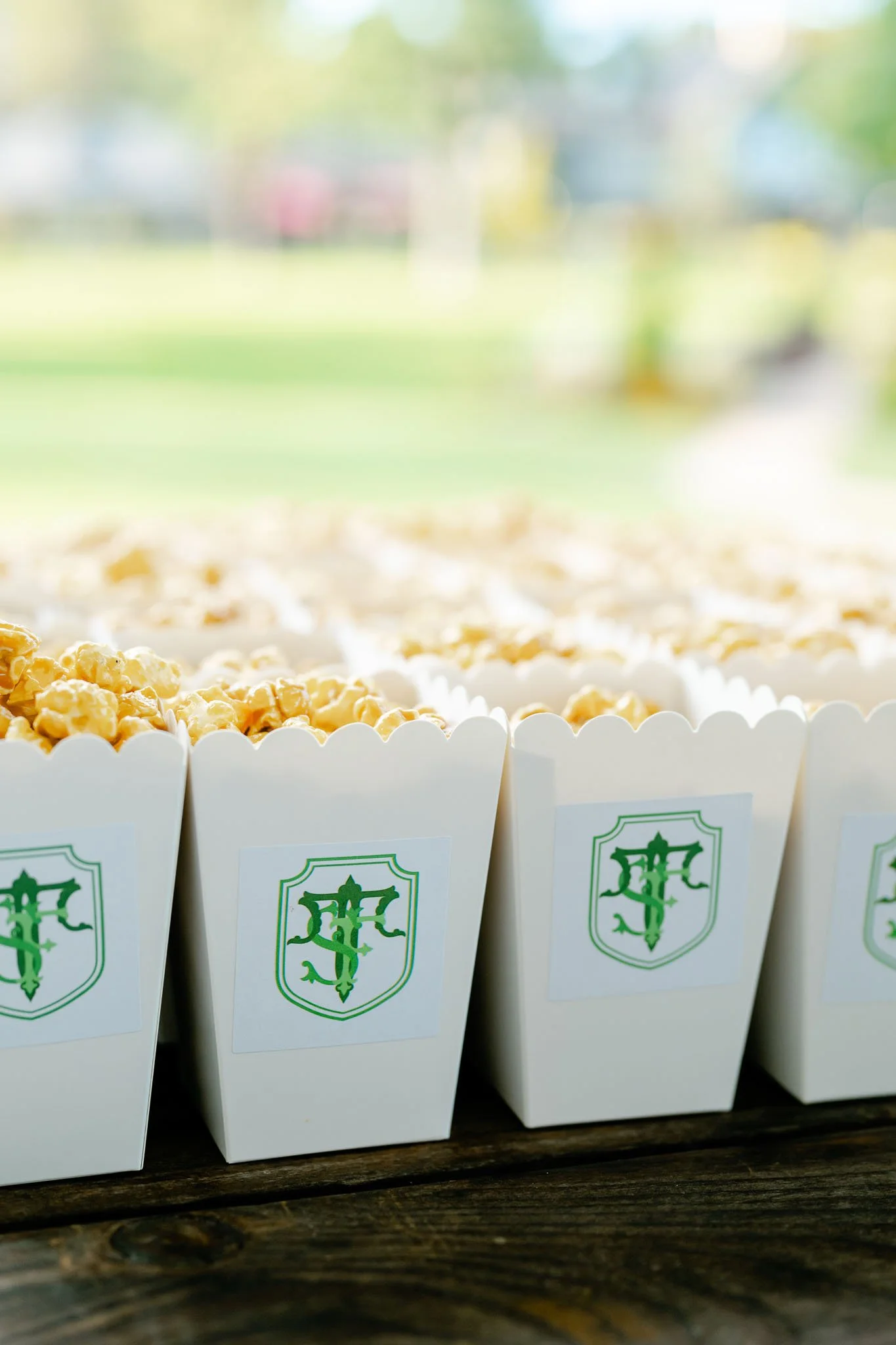 White popcorn boxes with a green emblem, filled with popcorn, displayed on a wooden surface outdoors with a blurred background of grass and trees.