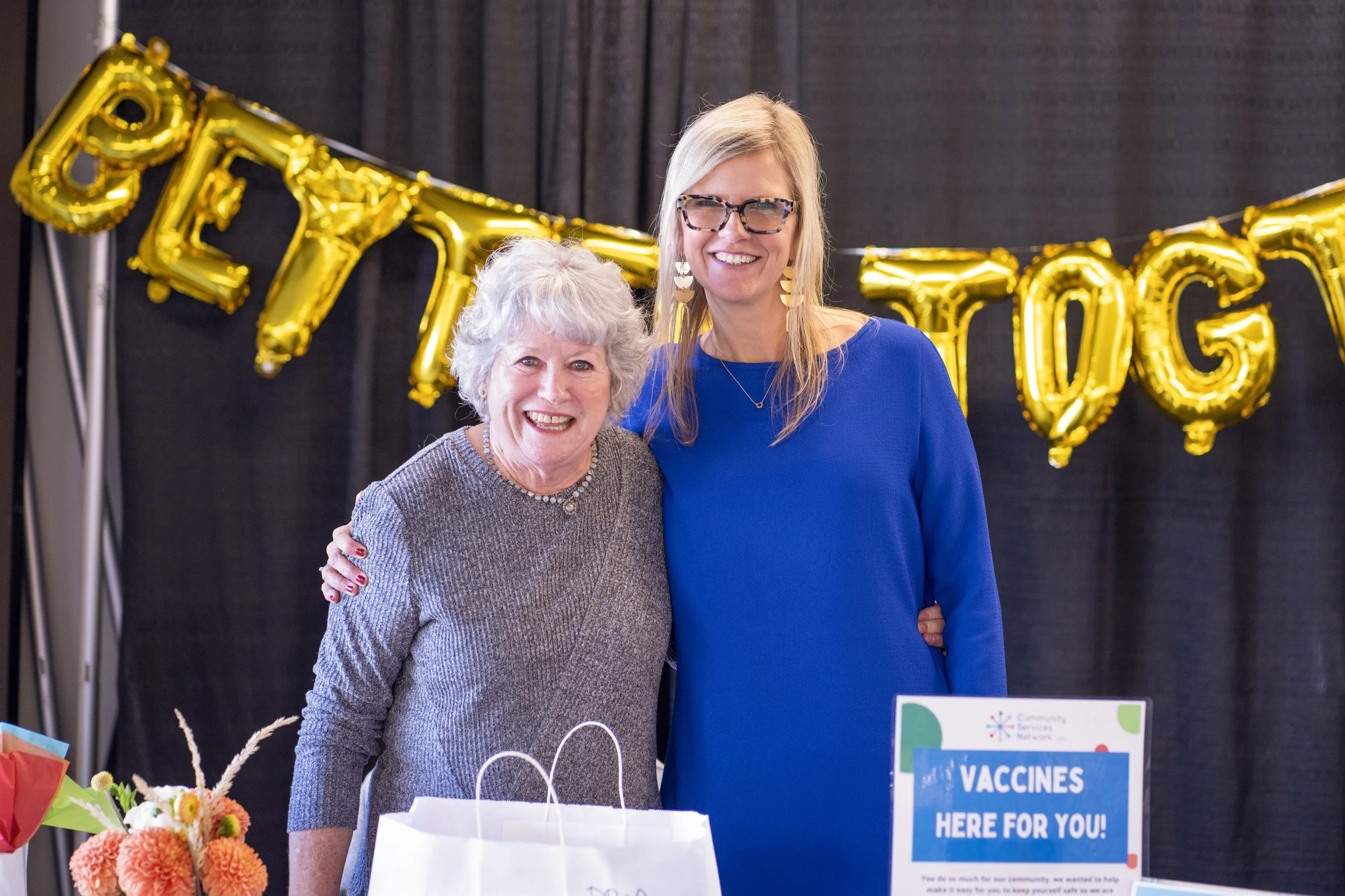  Two women posing together in front of gold 'Better Together' balloon letters and a Community Services Network vaccines sign at a CSN event. 