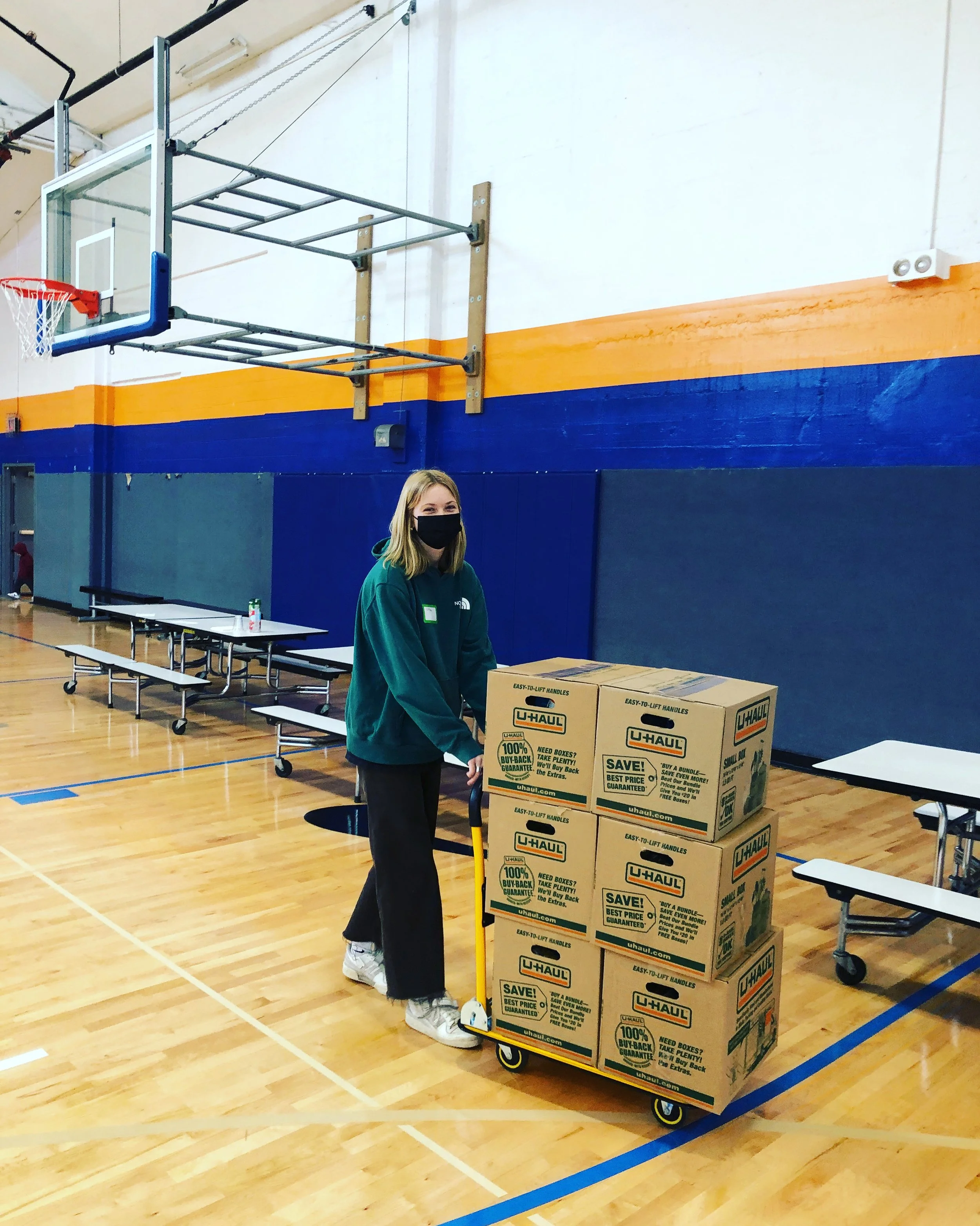  A young CSN volunteer in a green sweatshirt transporting supply boxes on a hand truck inside a school gymnasium to set up a community resource fair. 