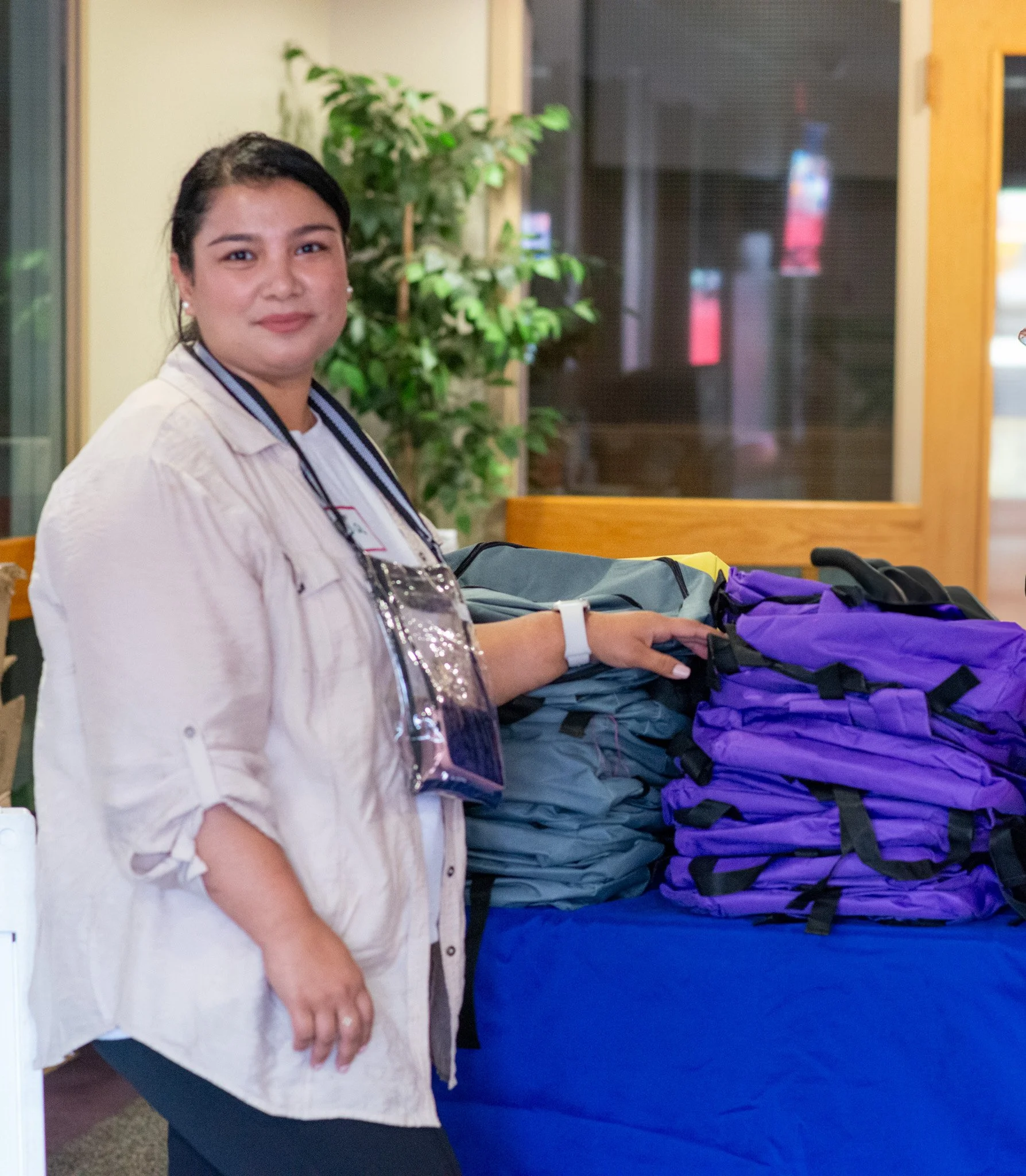  A smiling CSN volunteer standing beside a table stacked with purple and gray backpacks at a CSN community resource fair supply distribution event in Portland, Oregon. 