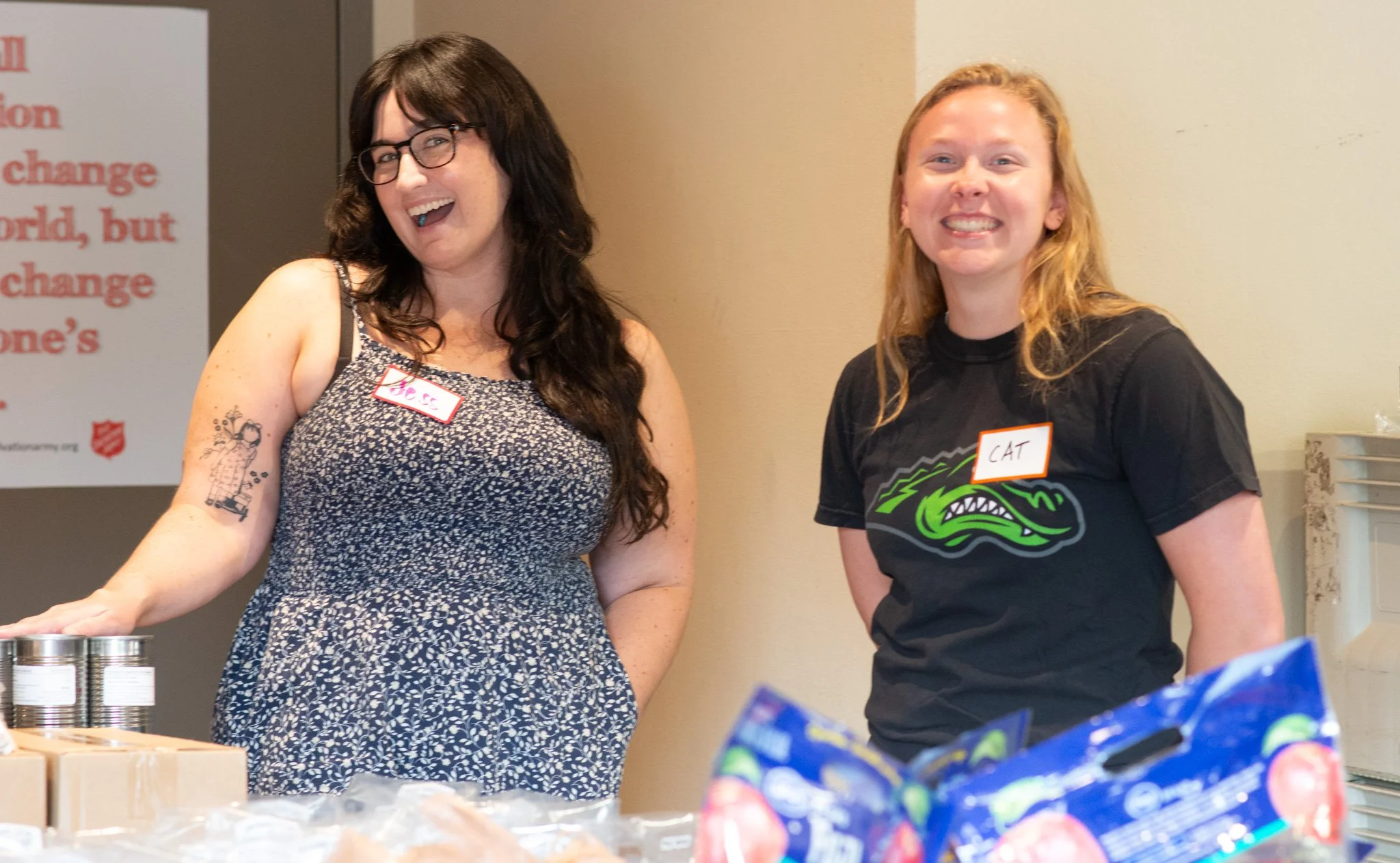  Two smiling CSN volunteers wearing name tags at a Salvation Army food distribution table with canned goods and packaged foods during a community resource event in Portland, Oregon. 