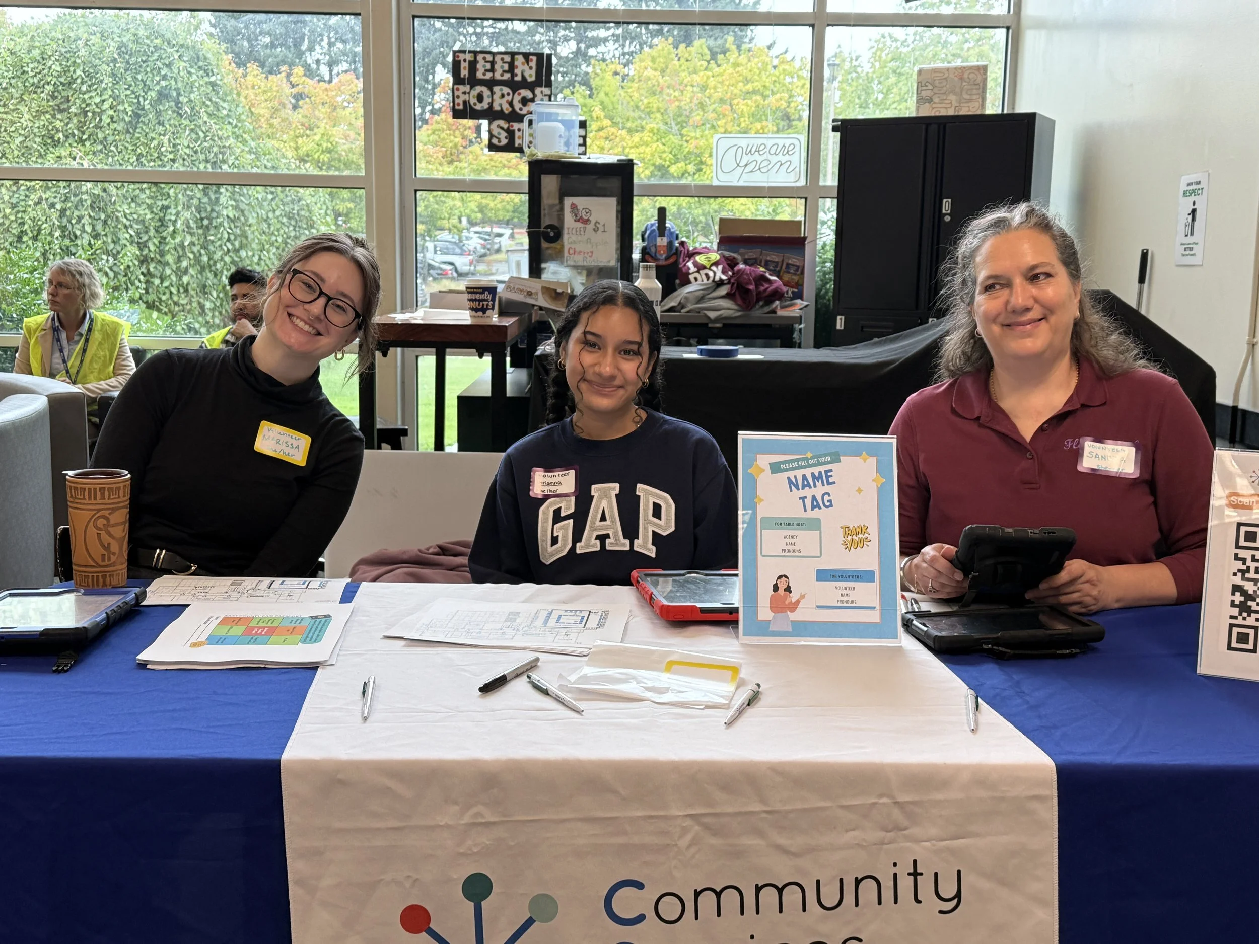  Three CSN volunteers smiling at a Community Services Network registration table at an indoor community resource fair in Portland, Oregon. 