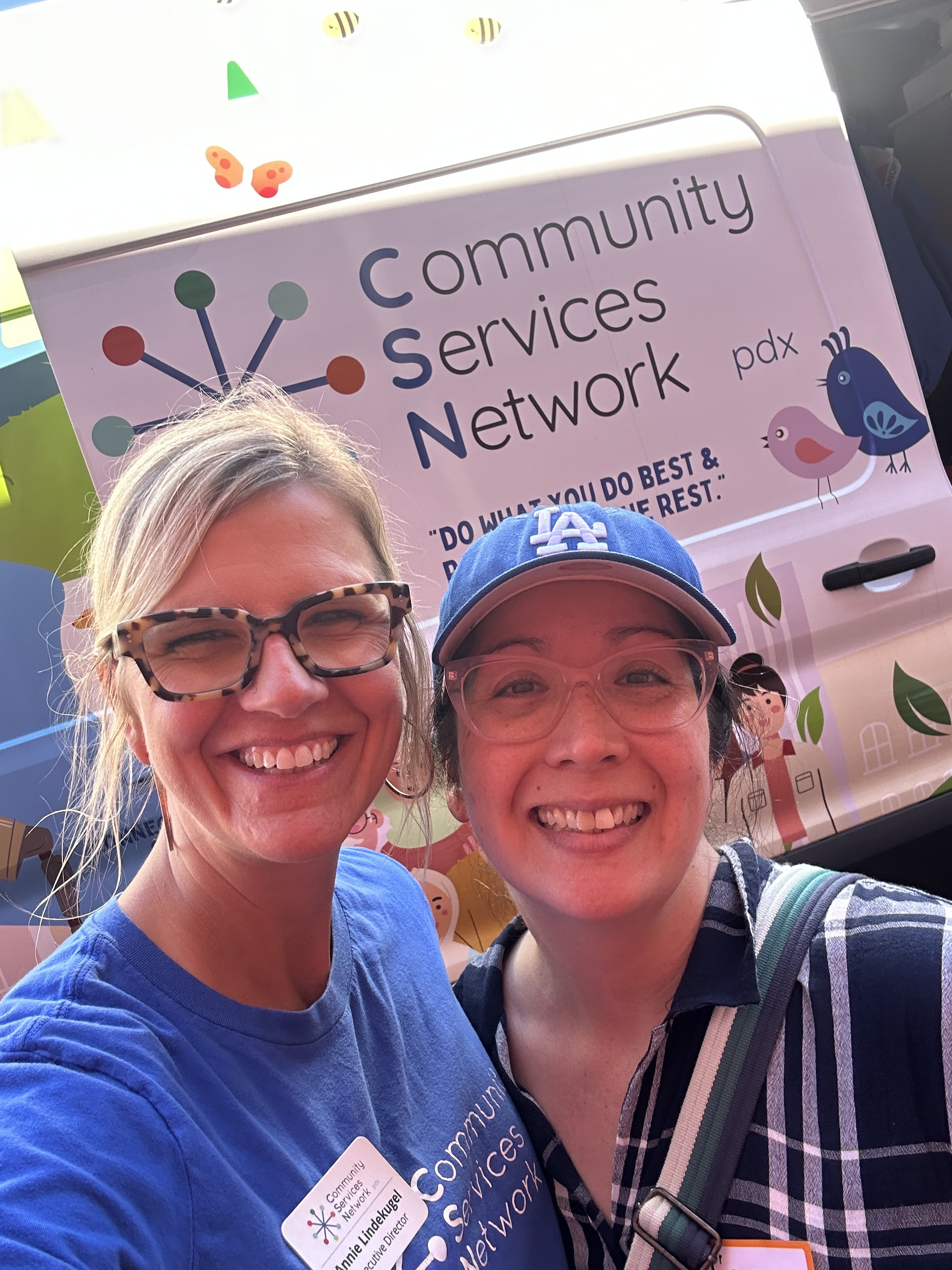  Two smiling Community Services Network PDX team members posing together in front of a CSN van at an outdoor community resource fair. 