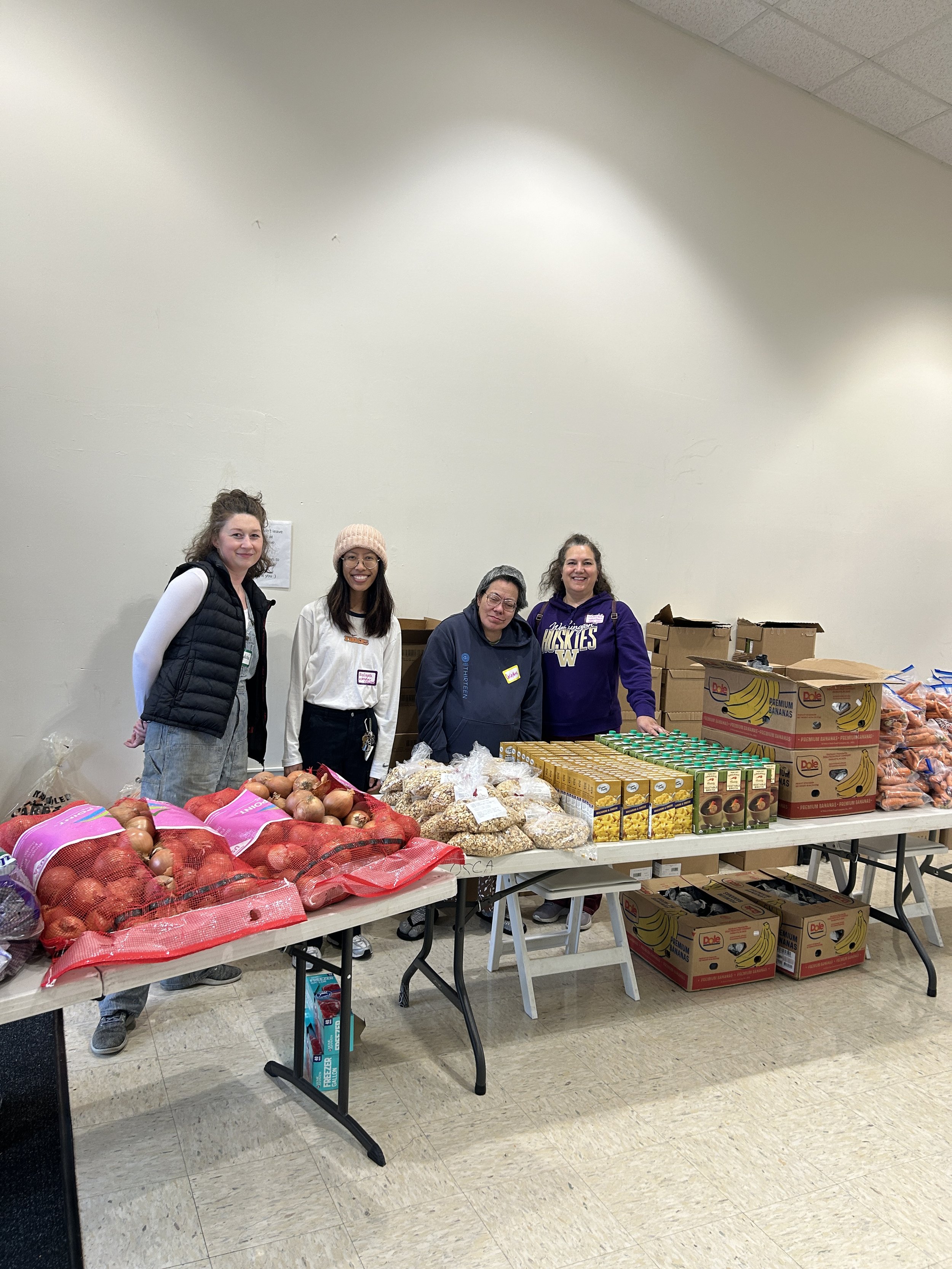  Three CSN volunteers standing behind a food distribution table stocked with fresh produce including onions, bananas, and other groceries at a community resource fair in Portland, Oregon. 