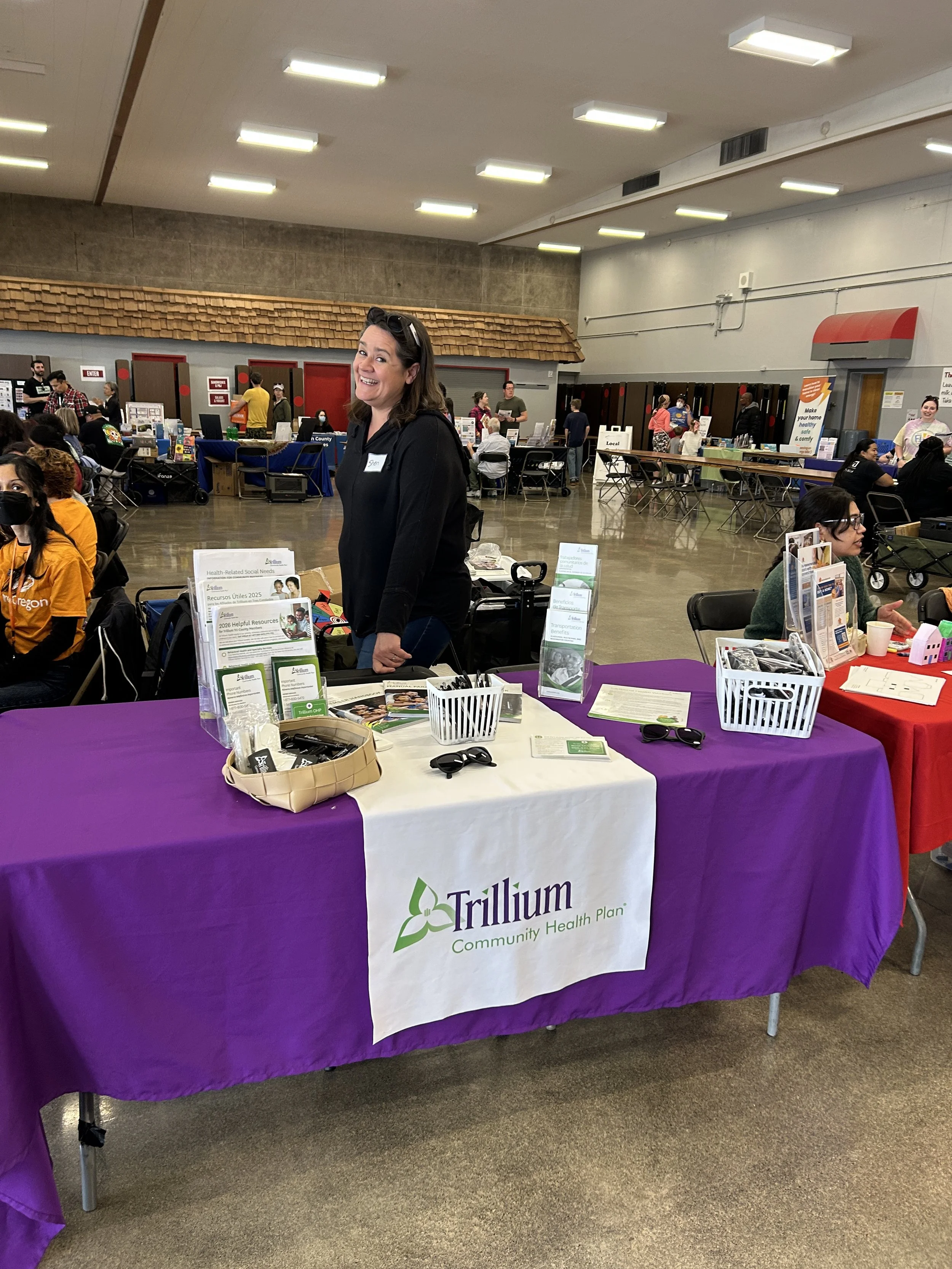  A Trillium Community Health Plan representative staffing a purple sponsor booth at an indoor CSN community resource fair in Portland, Oregon. 