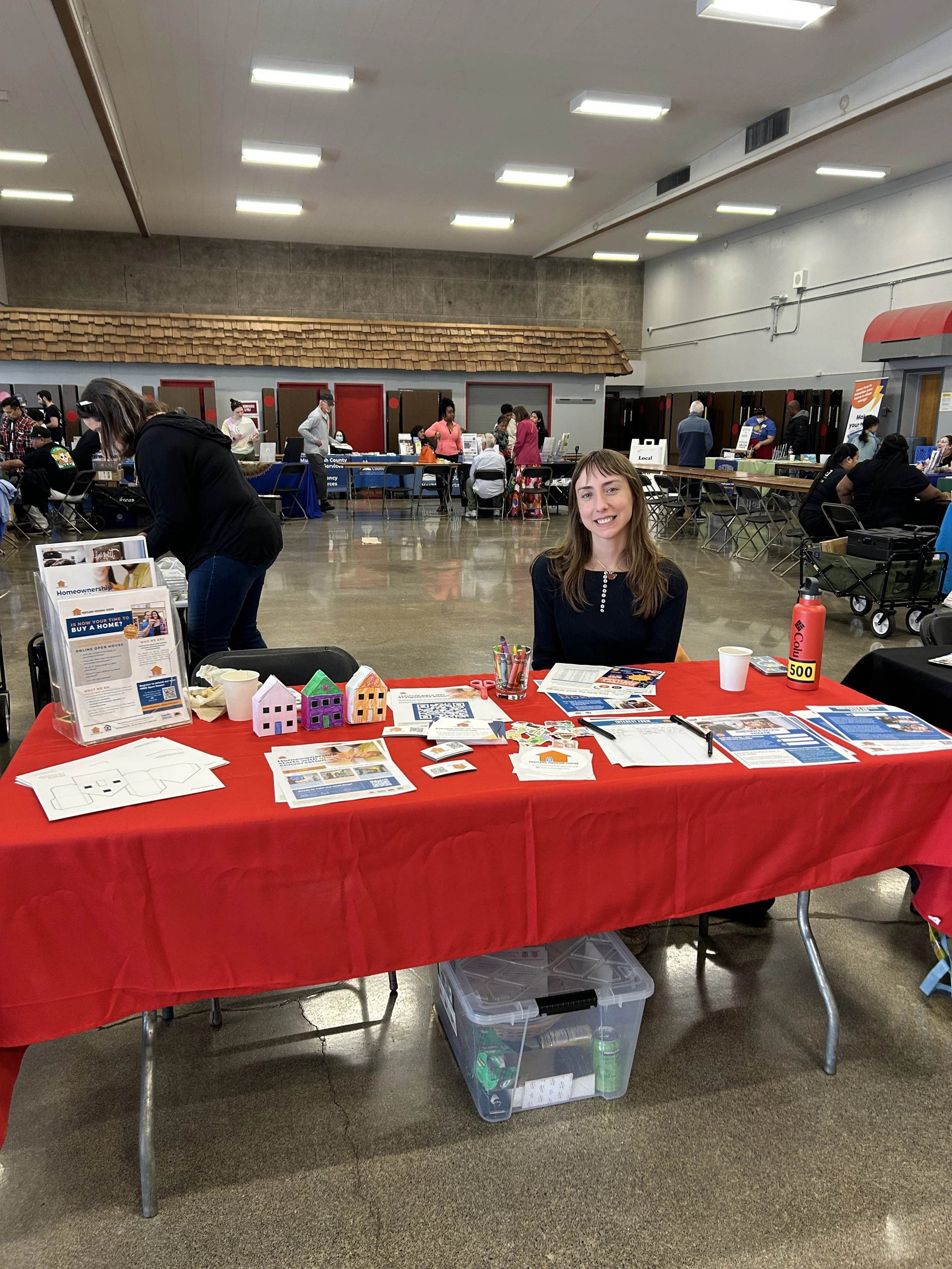  A sponsor representative staffing a red-draped resource table at an indoor CSN community resource fair in Portland, Oregon. 
