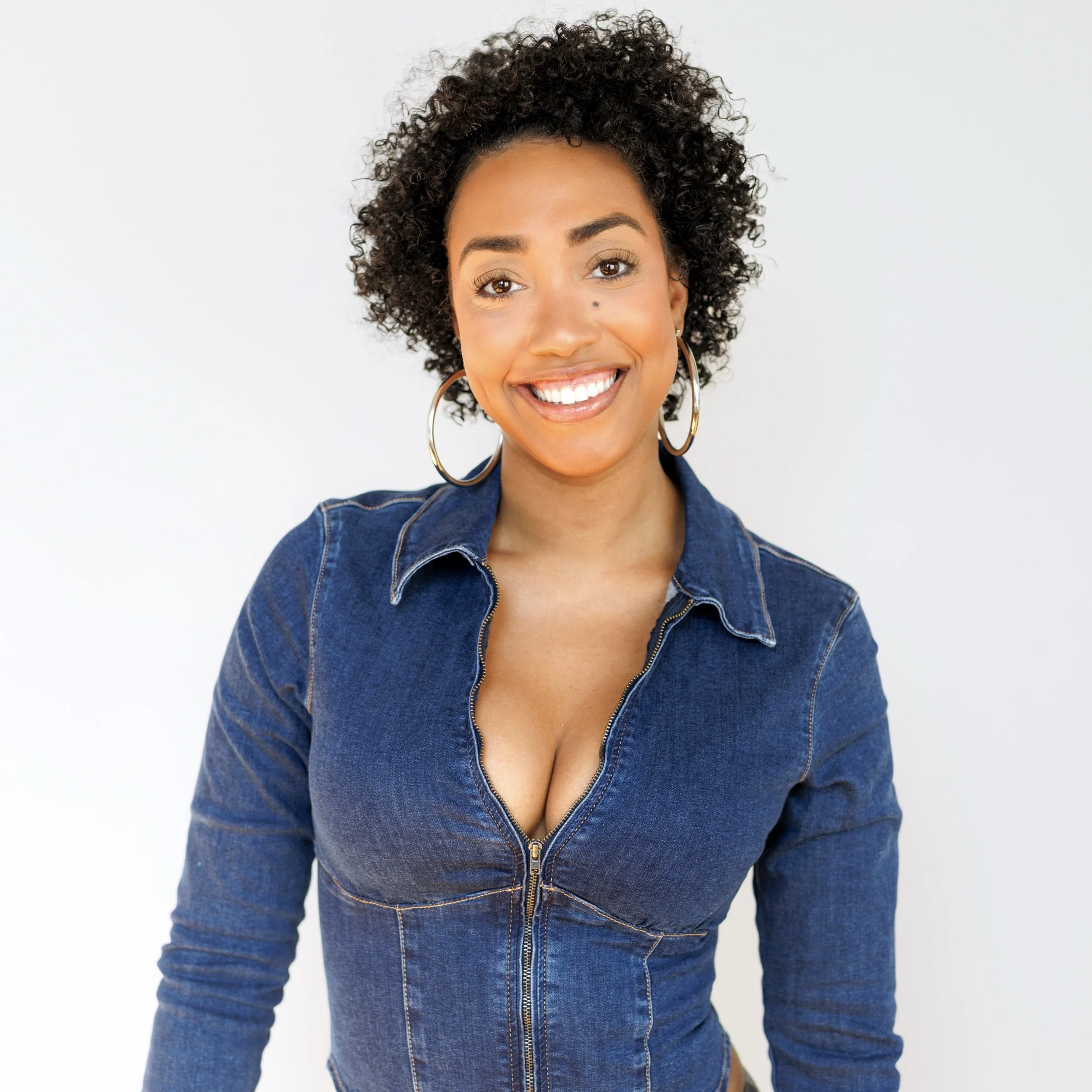 Smiling woman with curly hair, hoop earrings, wearing a denim jacket, against a plain light background.