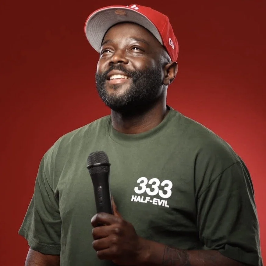 A man with a beard, wearing a red cap and a green T-shirt with "333 HALF-EVIL" written on it, holds a microphone and smiles against a red background.