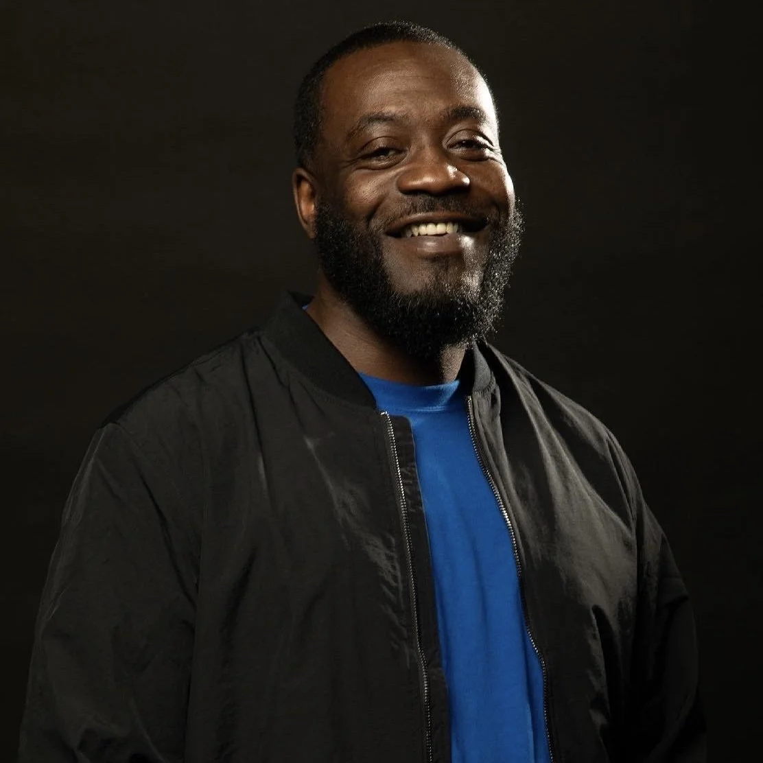 A smiling man with a beard wearing a black jacket over a blue shirt, standing against a dark background.