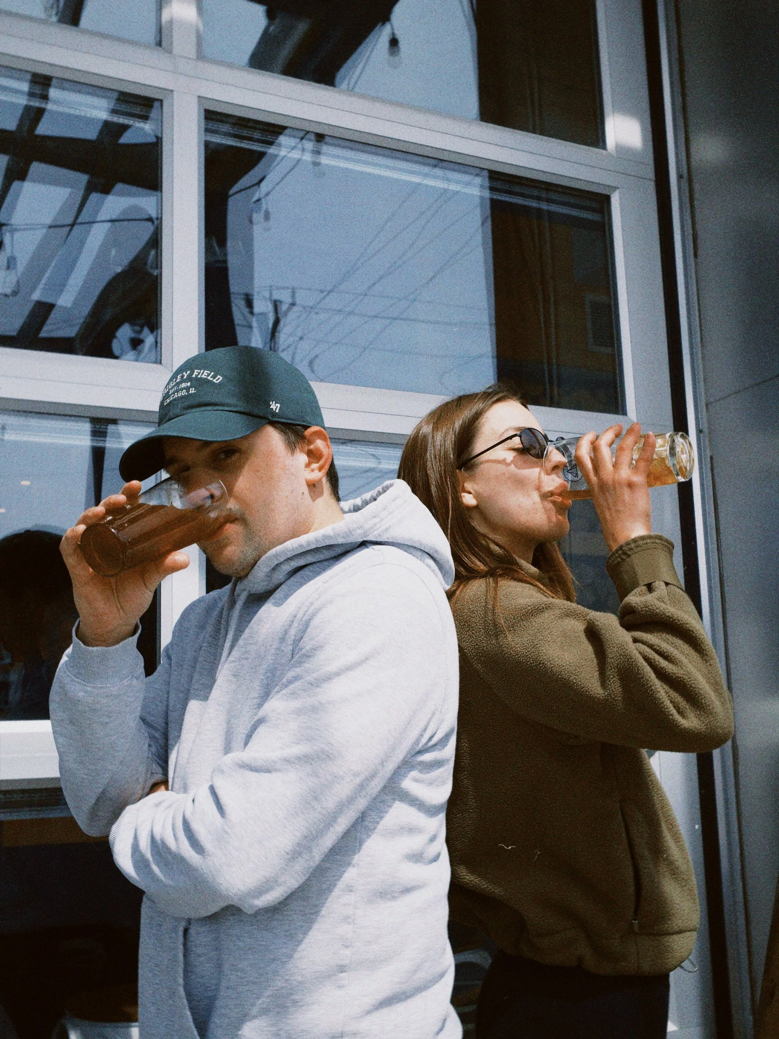 Two young adults, a man and a woman, standing outside a building with glass windows, both drinking from bottles of what appears to be dark soda.