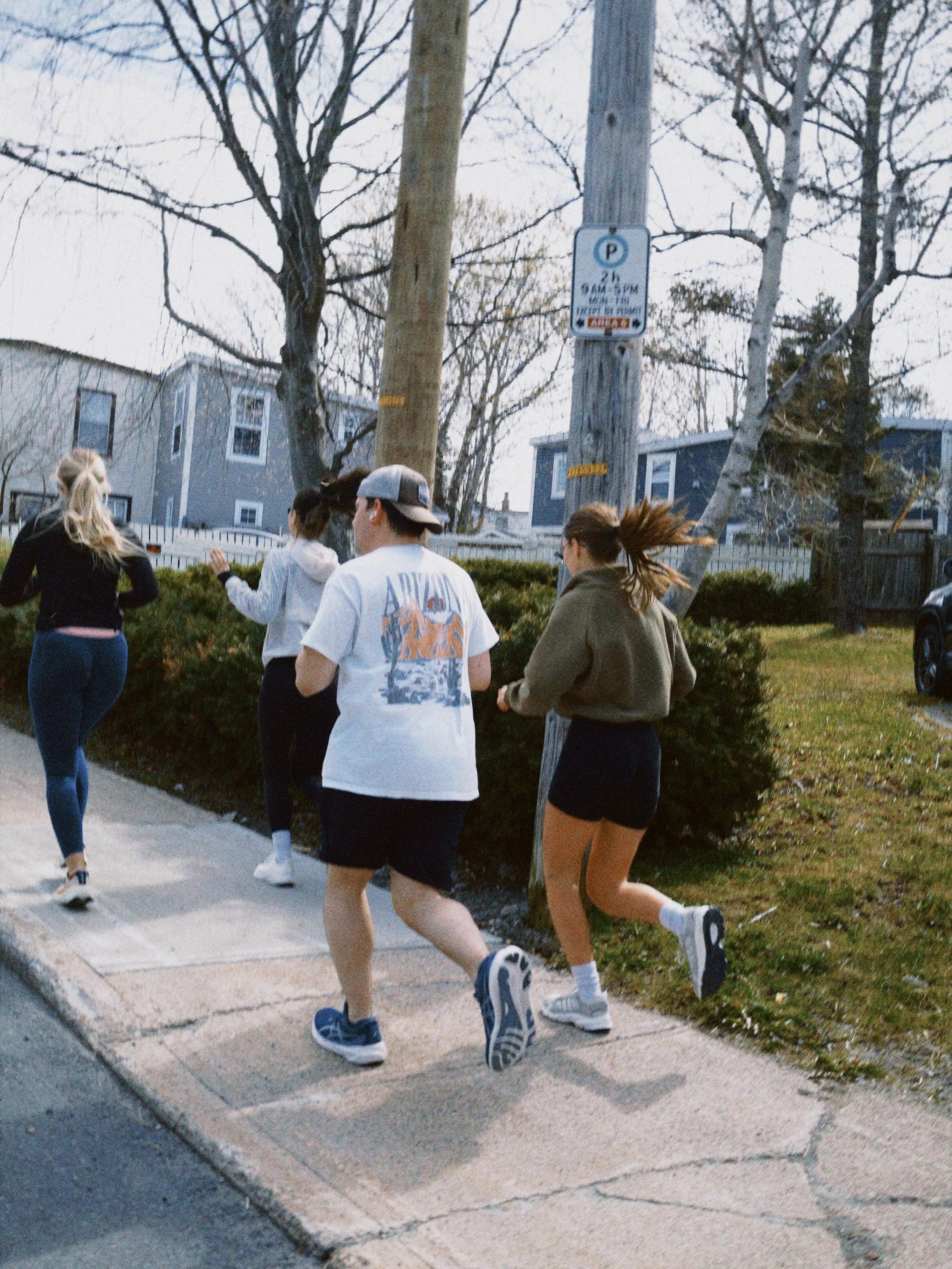 Four young people jogging on a sidewalk in a residential neighborhood with trees, houses, and a parking sign in the background.