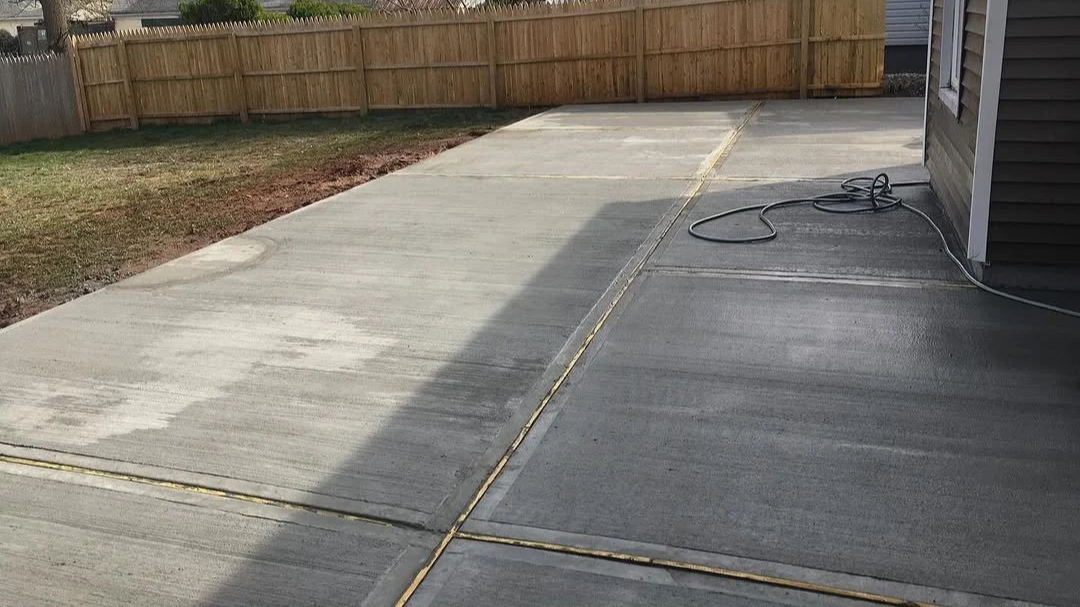Freshly cleaned concrete sidewalk and driveway next to a house, with a wooden fence in the background and a garden hose coiled on the side of the house.