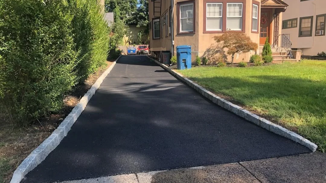 Freshly paved black asphalt driveway with white brick borders, leading to a house with a front porch and steps. Green grass lawn and bushes on the left, several blue trash bins at the end of the driveway.