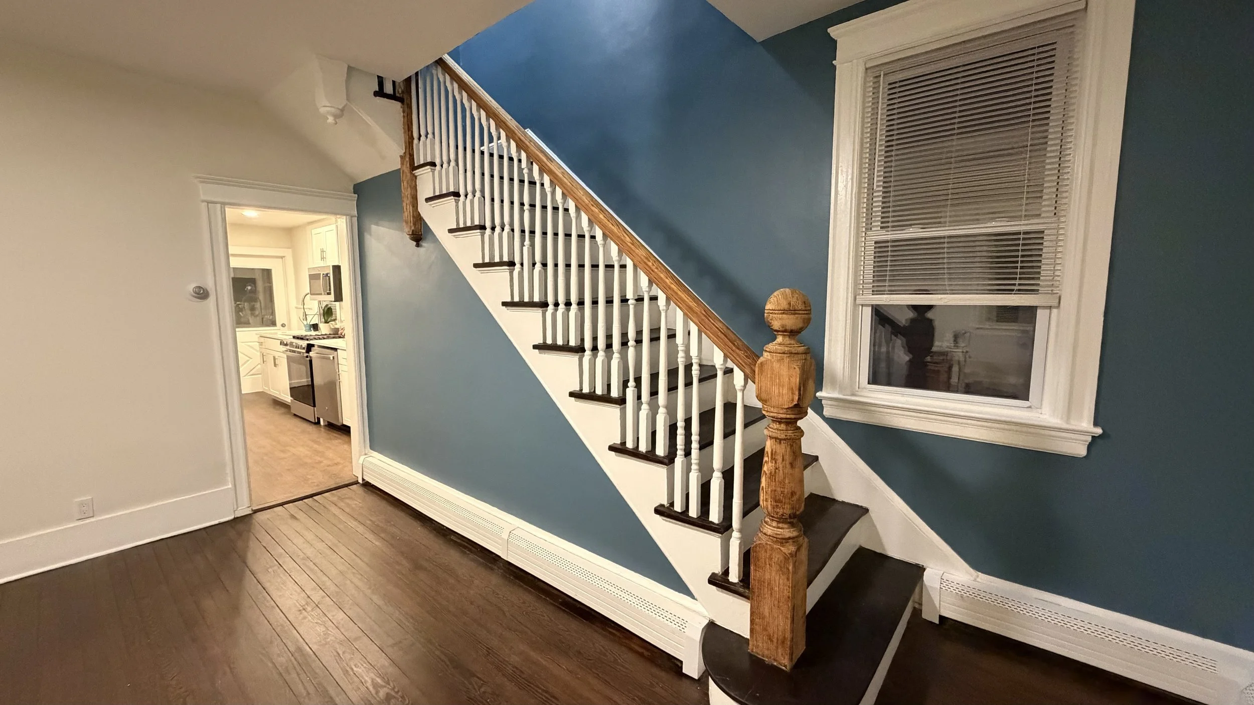 Interior of a home showing a staircase with white spindles and wooden handrail, blue accent wall, window with blinds, and doorway leading to the kitchen.