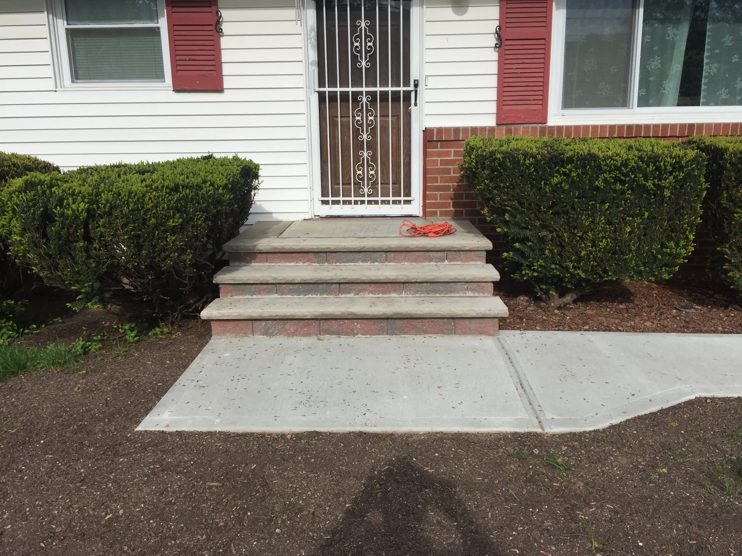 Concrete steps leading up to a front door with a security screen, flanked by bushes, with a sidewalk in front.