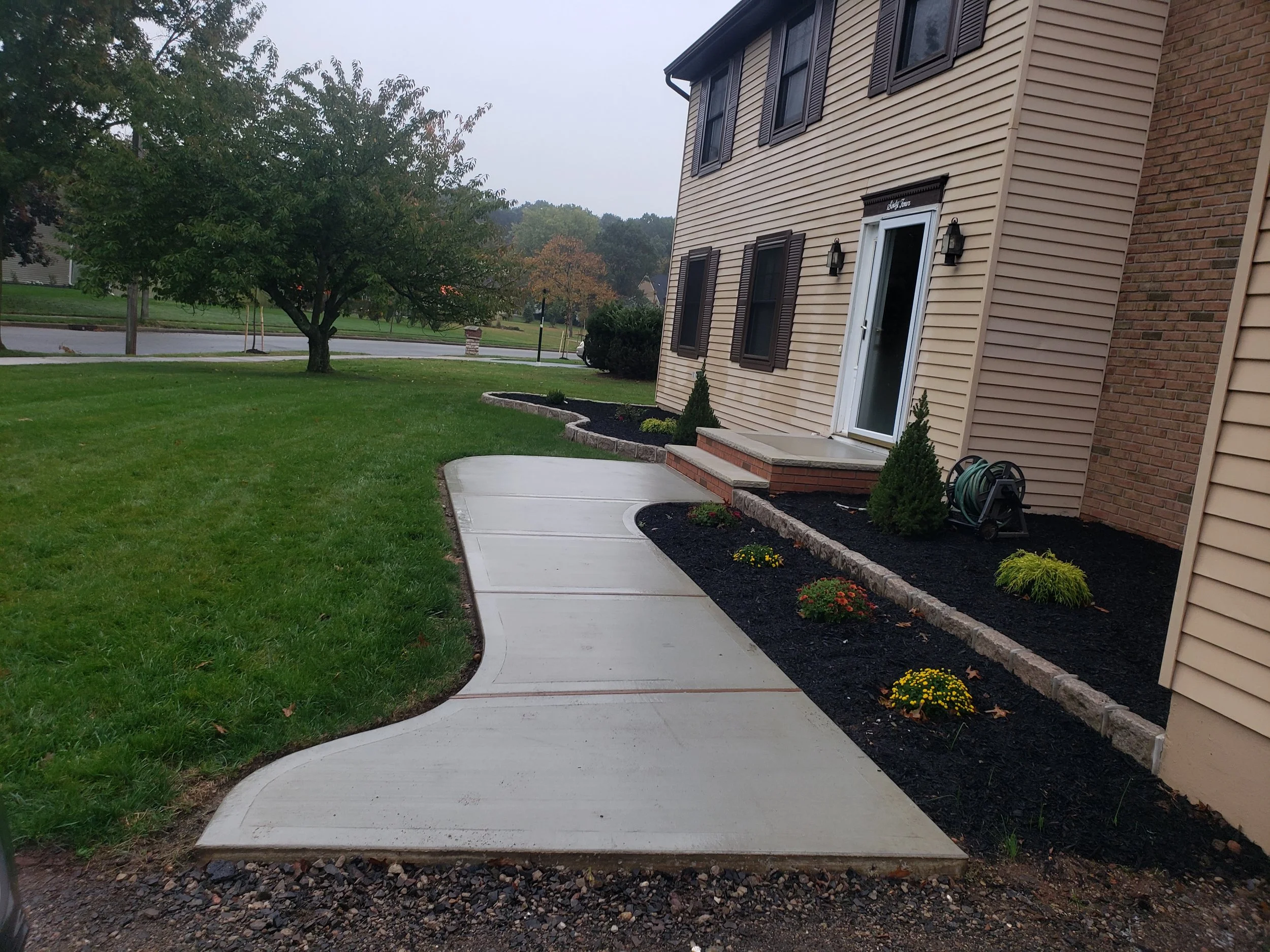 Concrete sidewalk curves in front of a beige house with brown shutters, flower beds with small bushes and yellow flowers, and a green lawn with a large tree in the background.