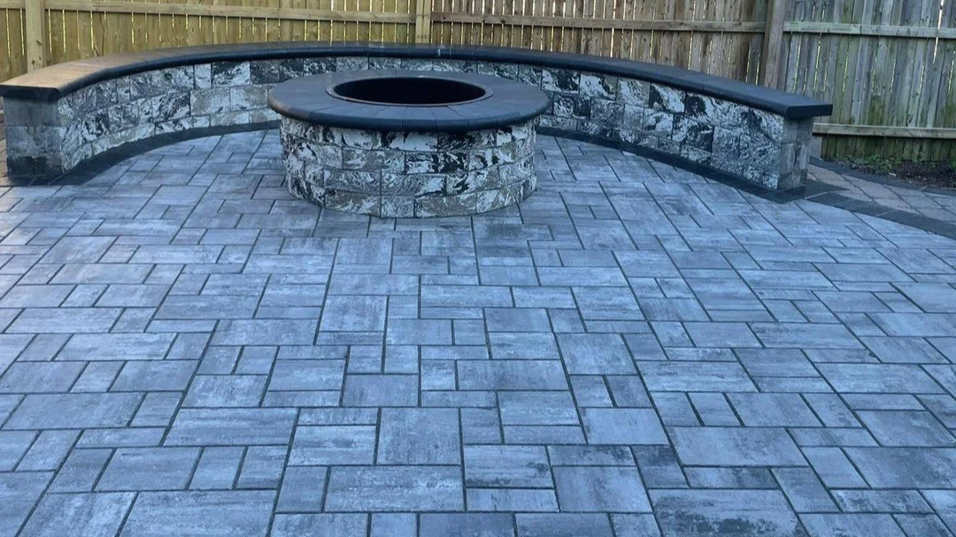 Round stone fire pit surrounded by a curved stone bench on a paved patio with a wooden fence in the background.