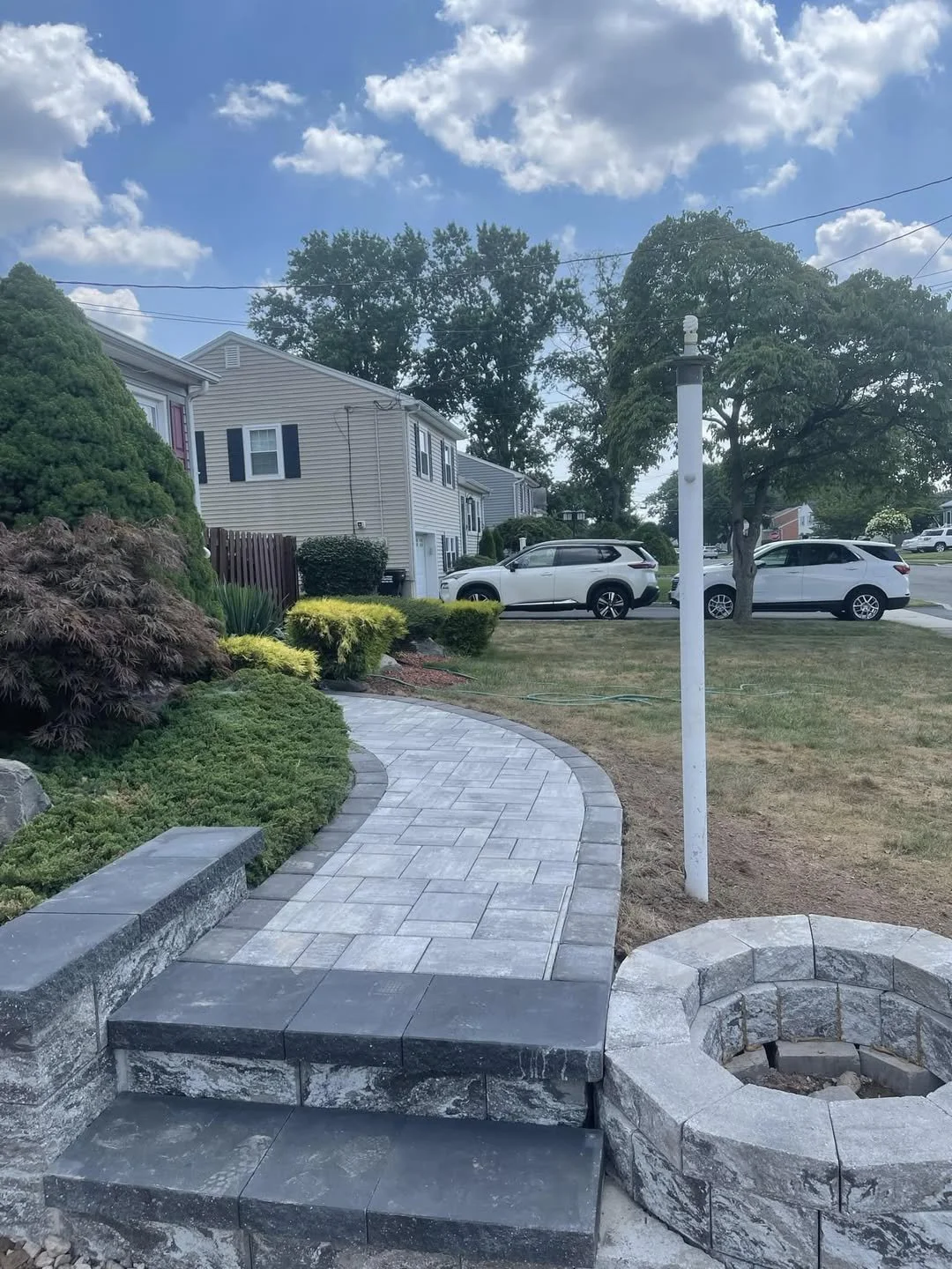 A newly paved, curved gray stone walkway in front of a suburban house, with small plants and bushes along the sides, a white patio post, and two white cars parked on the street under a partly cloudy sky.