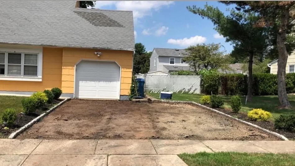 A house with a front yard undergoing landscaping, with the driveway cleared and bordered by small bushes, and a sidewalk in the foreground.