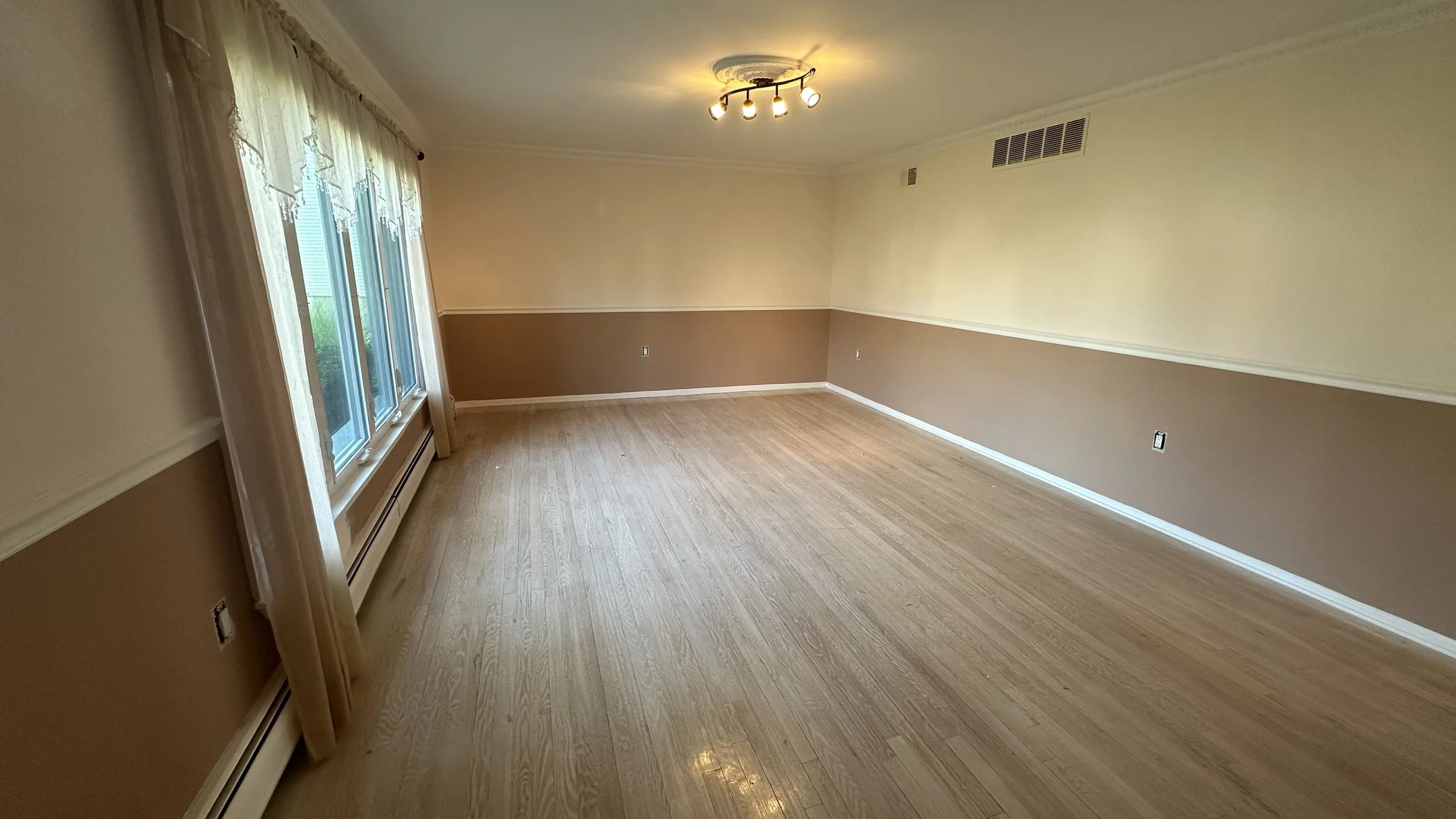 Empty living room with beige and light brown walls, large window with curtains, light wood flooring, and ceiling light fixture.