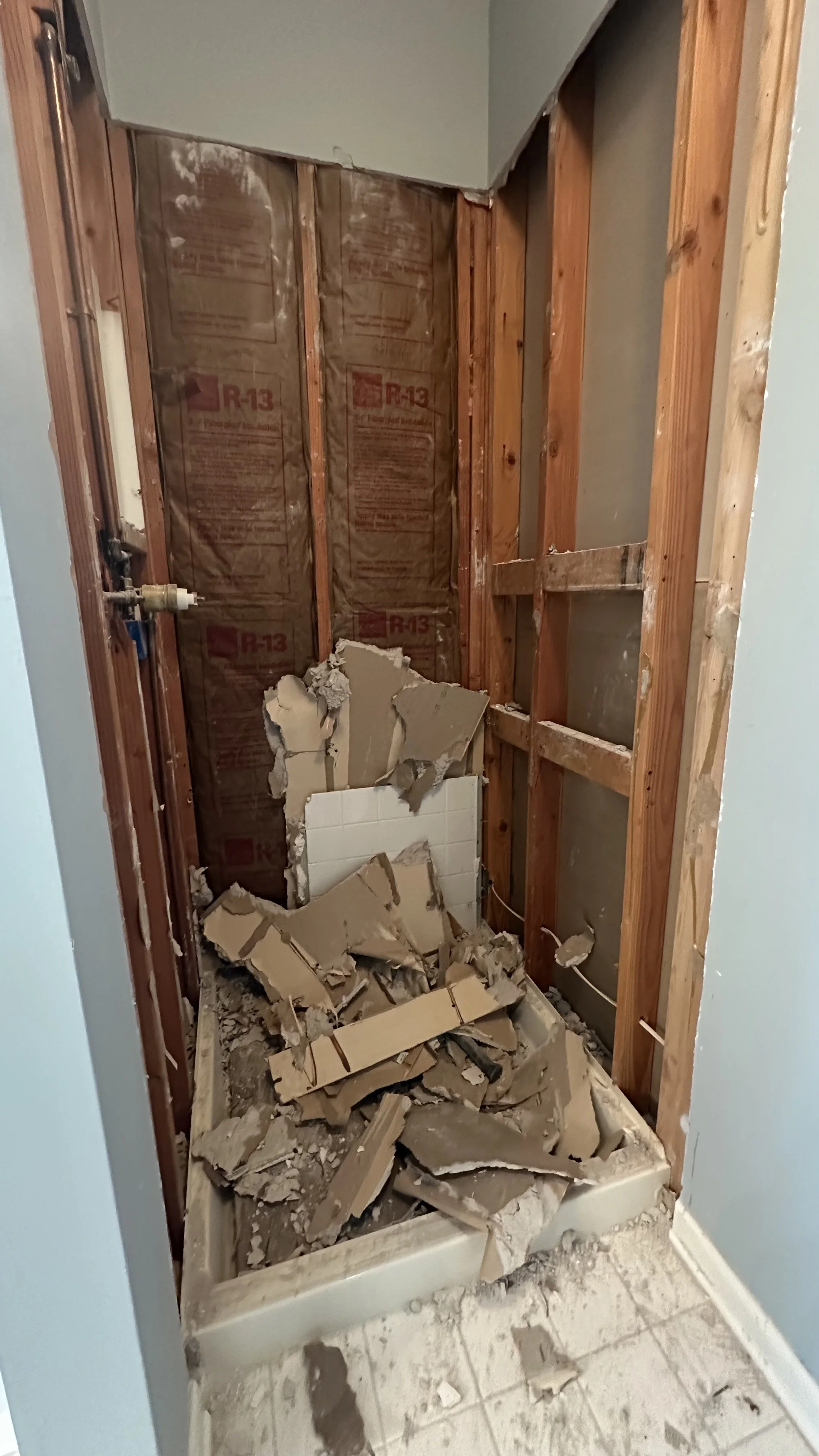A bathroom under renovation with exposed studs, insulation, broken tiles, and debris on the floor.