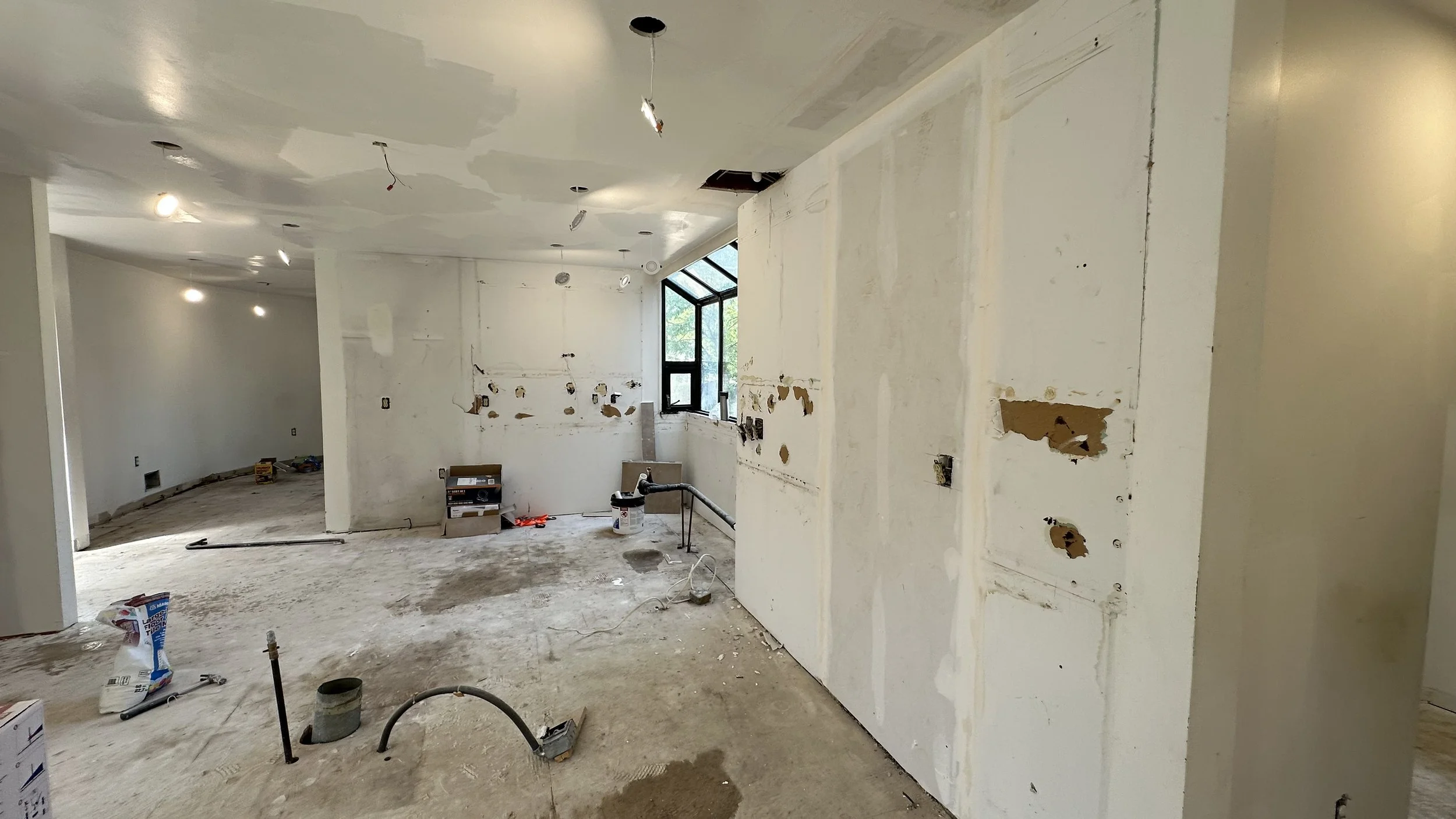 Interior of a room under construction with exposed drywall, ceiling lights, and construction tools and materials on the floor.