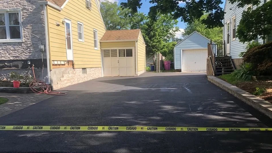 Freshly paved black driveway with caution tape across the front, leading to yellow and white garages and surrounded by green trees and plants.