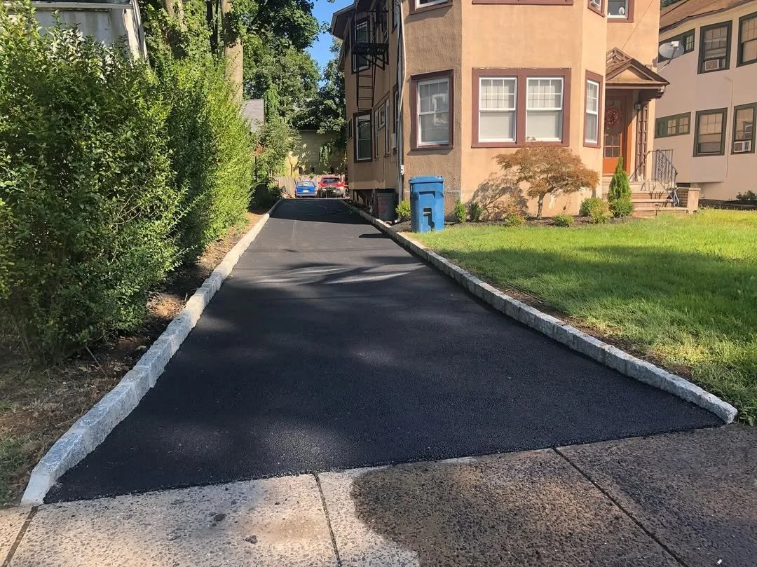 Recently paved driveway with fresh black asphalt, edged with light-colored bricks, leading to a residential building and parked cars in the background.