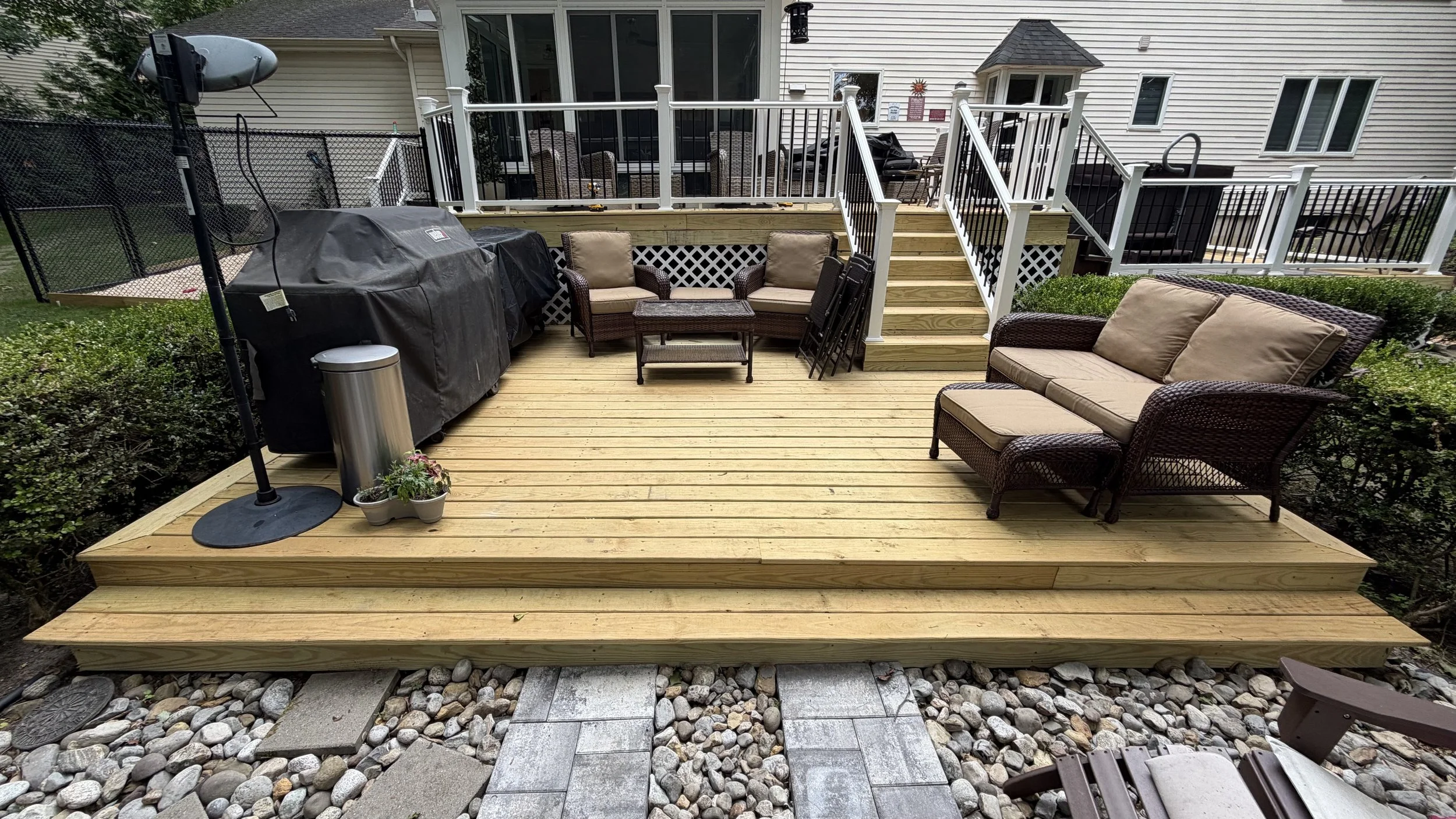 A wooden deck with outdoor furniture including a sofa, chairs, and a small table. There are stairs leading up to an elevated section with more chairs and a grill covered with a black cover. A satellite dish and some potted plants are also visible.