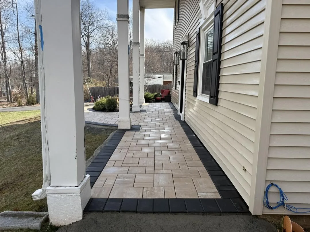 View of a covered porch with beige and black paving stones, white posts, and beige siding, with a backyard containing trees and a lawn.