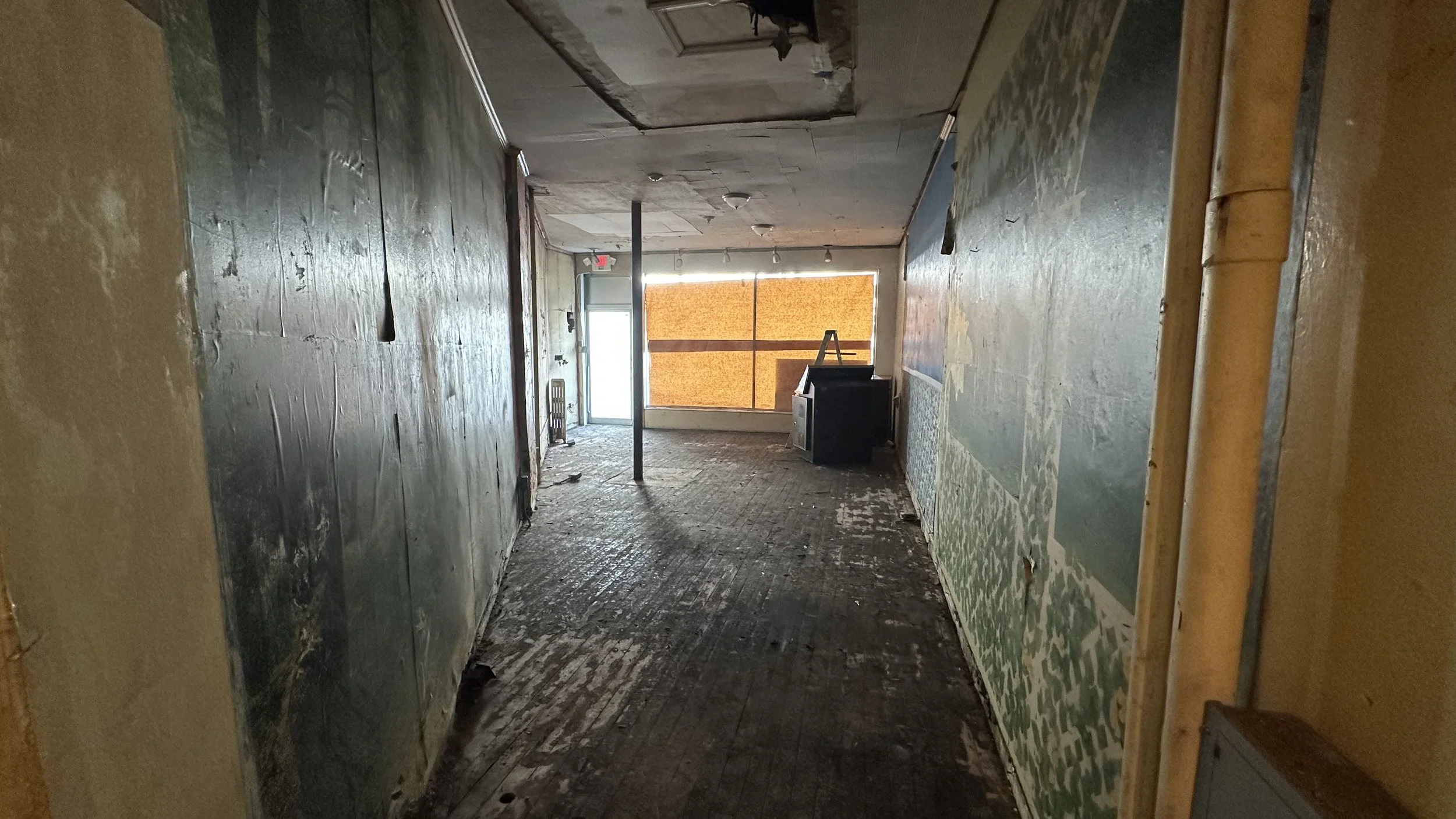Interior of a dilapidated building with peeling paint, worn flooring, and boarded-up windows, leading to an exit door.