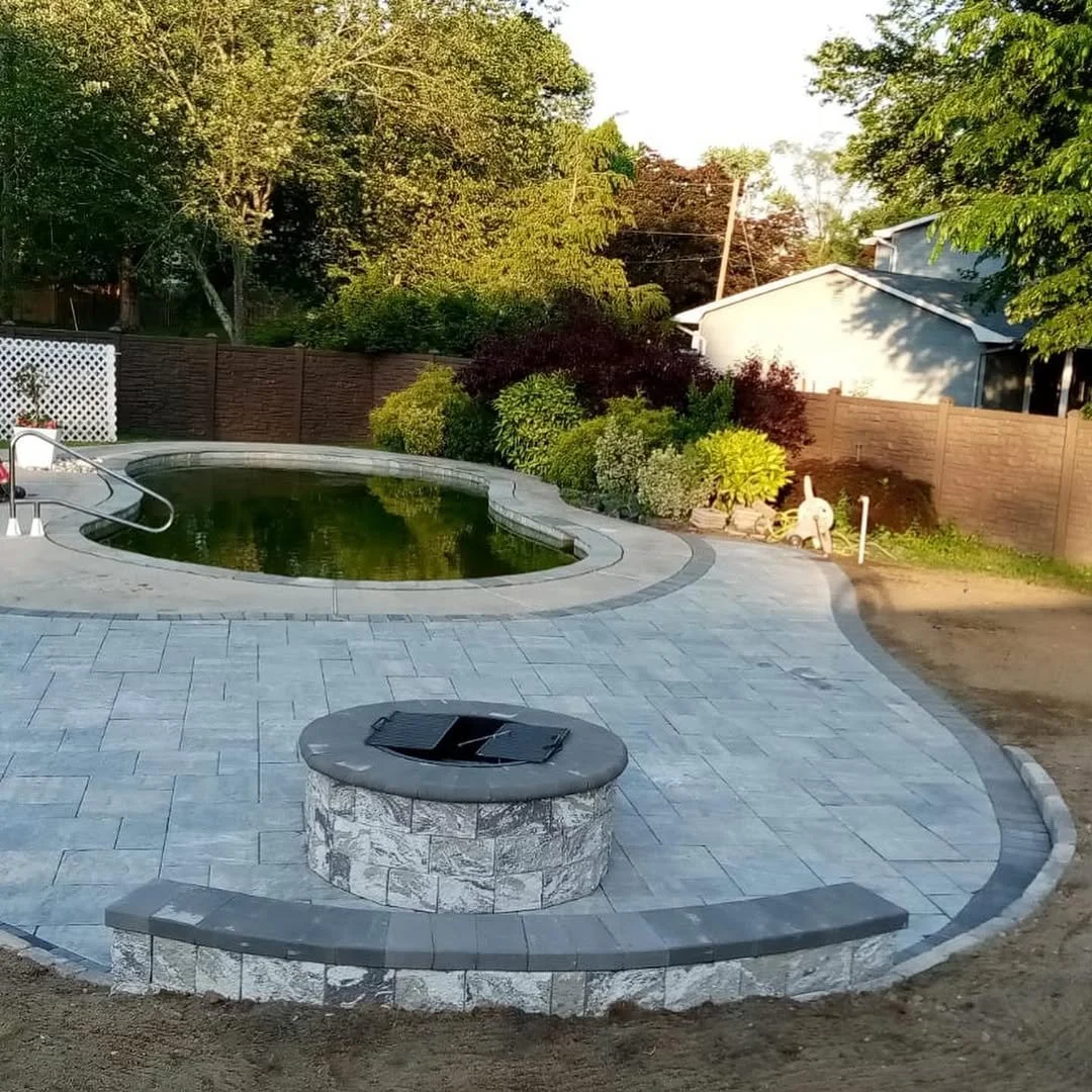 A backyard with a pool, surrounding patio, a fire pit with a grill on top, and greenery including trees and bushes, with a house and fence in the background.