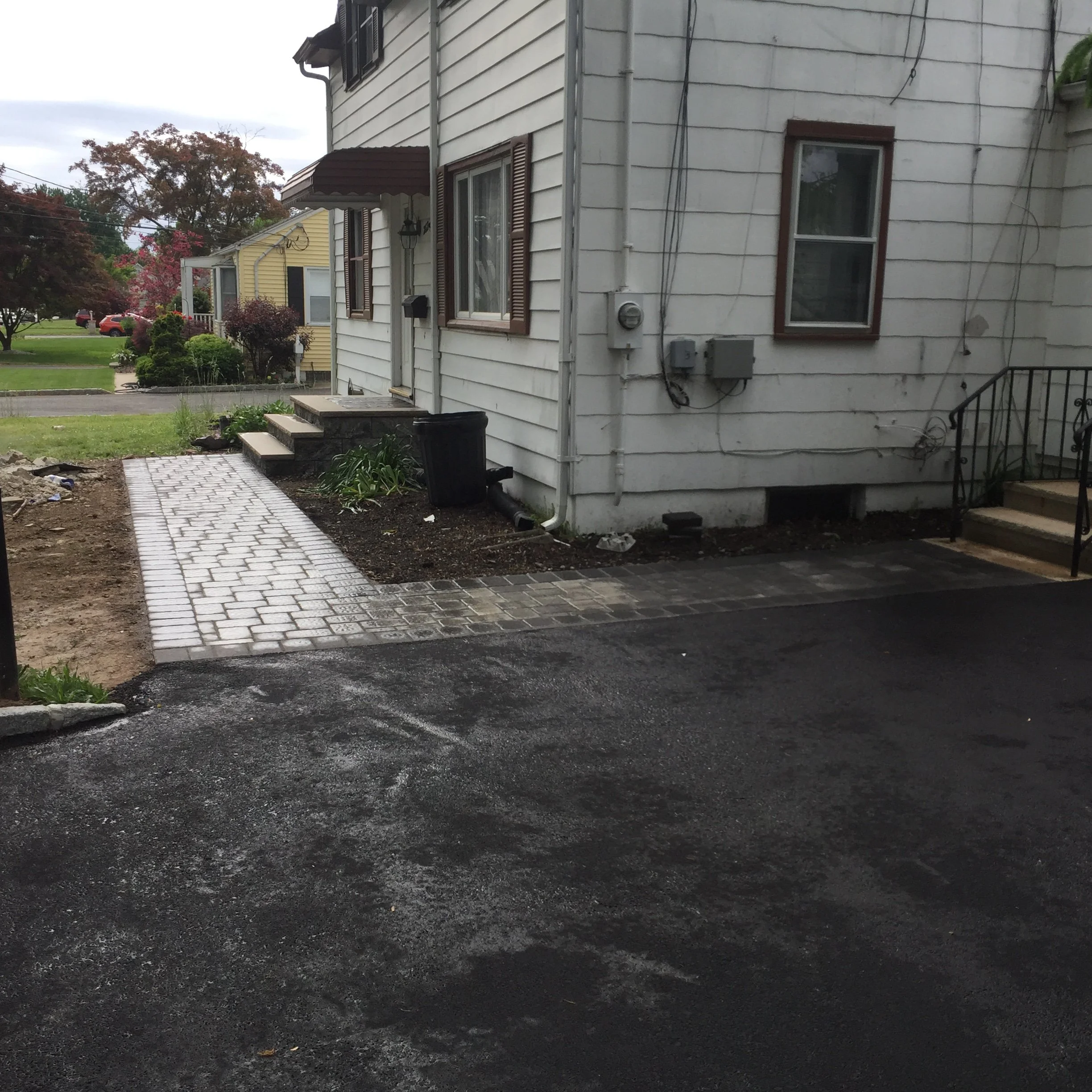 A newly paved asphalt driveway leading to a house with a small brick pathway at the entrance. There are steps with a black metal railing on the right side, and a white house with brown window shutters. A small garden bed is on the left side of the pa