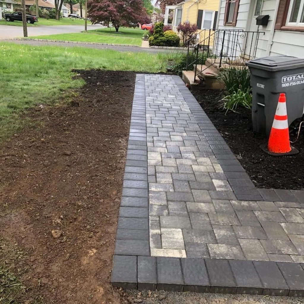 Newly paved brick walkway next to a house with steps, a railing, a trash bin, and a traffic cone, with a lawn and trees in the background.
