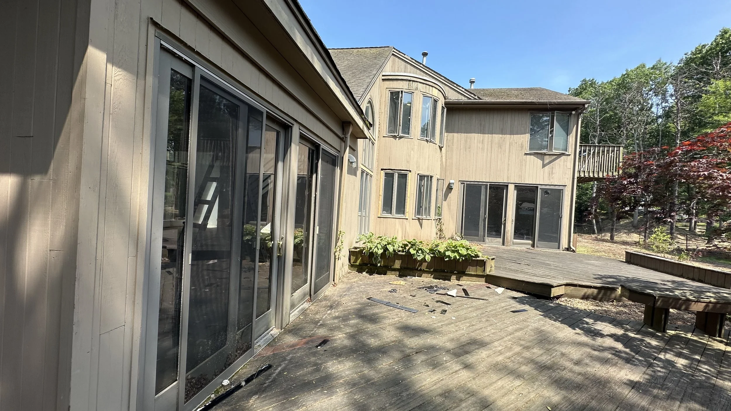 Backyard deck with sliding glass doors, a raised garden bed with plants, and outside trees with green and red foliage under a clear blue sky.
