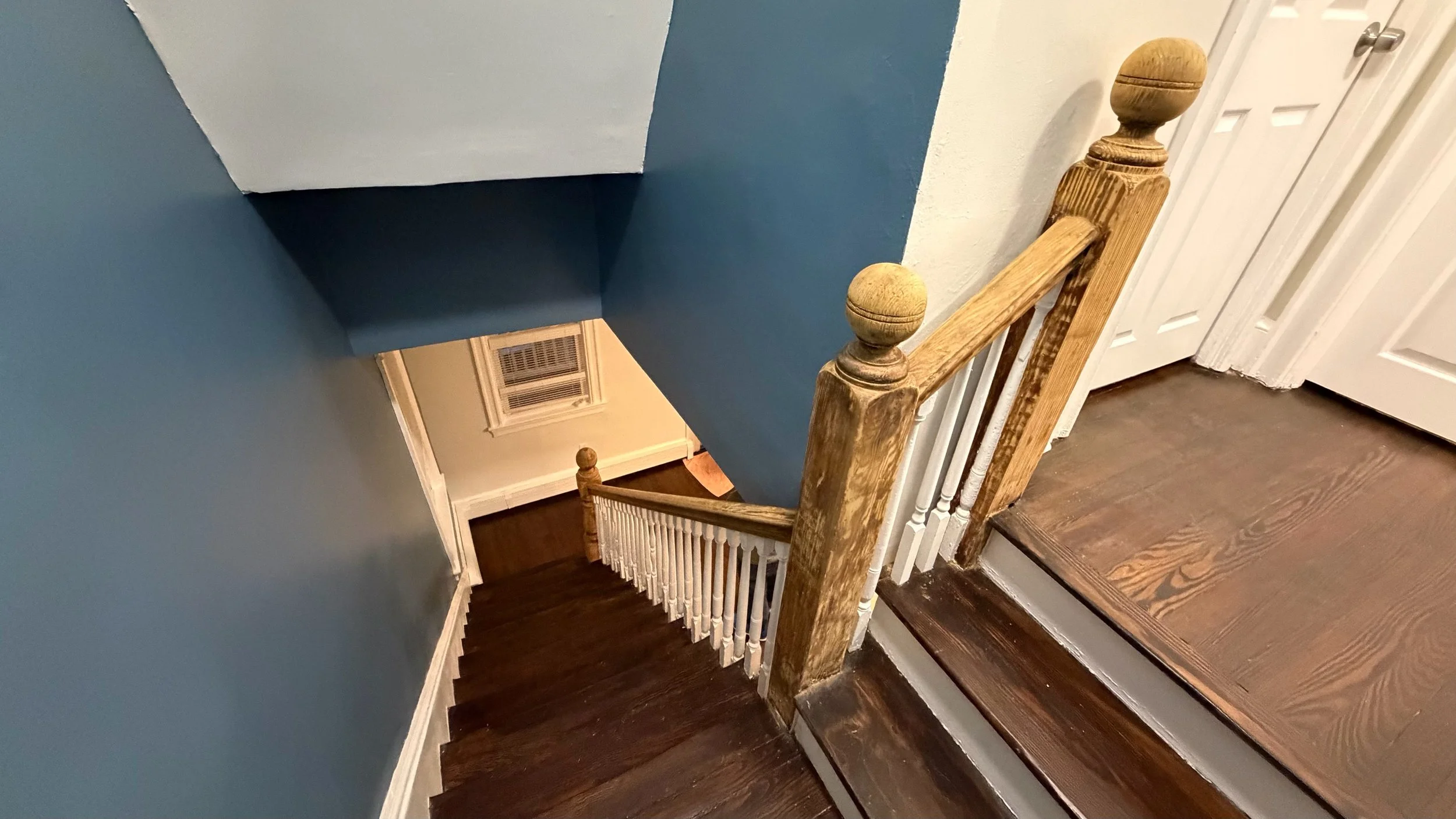 A wooden staircase with dark brown steps, white spindles, and a wooden handrail with a newel post, leading down to a landing. The walls are painted in light blue and white, with a white door visible at the top of the stairs and a window with an air vent on the wall near the bottom.