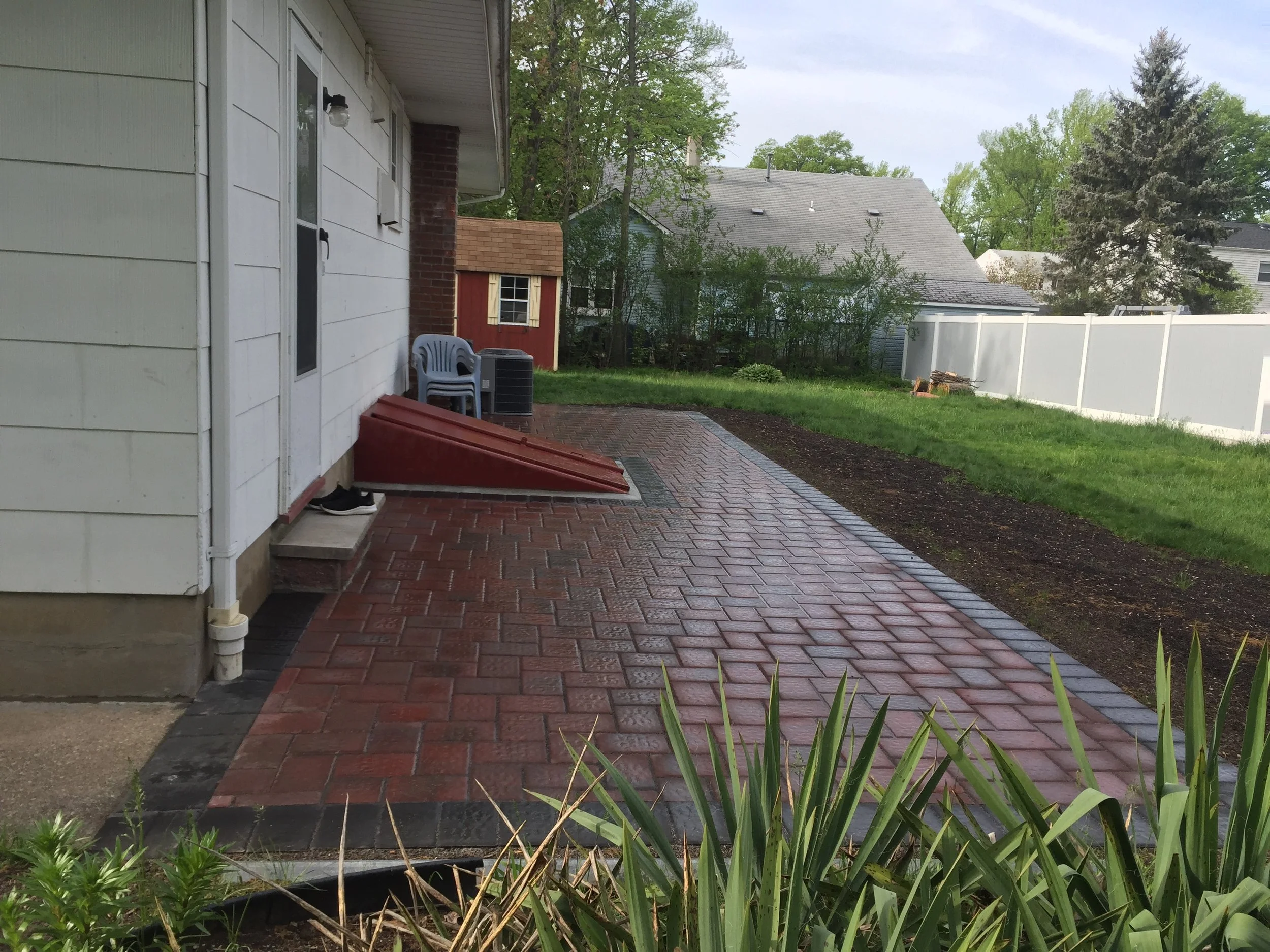 Backyard with brick patio, white house wall, plastic chairs, small red garden shed, and a white privacy fence. Green grass, trees, and a cloudy sky in the background.