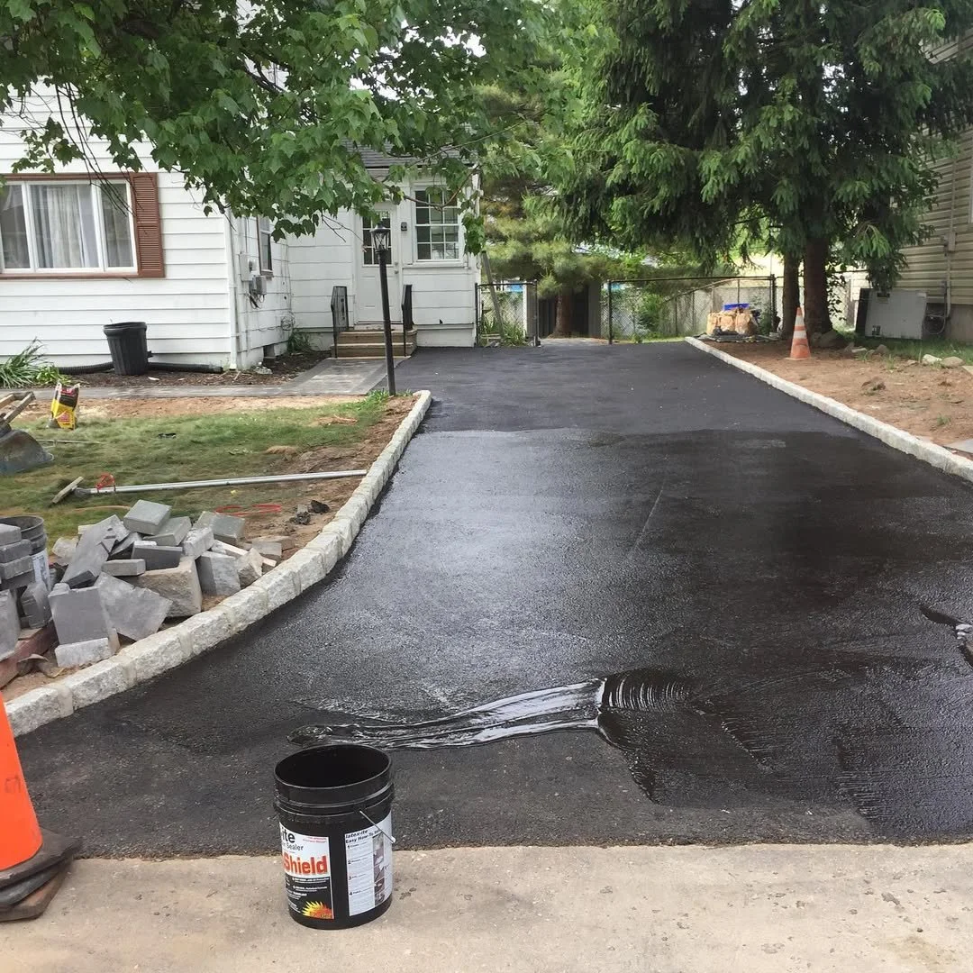 Freshly paved asphalt driveway with a bucket of black sealant, construction tools, and a pile of bricks nearby.